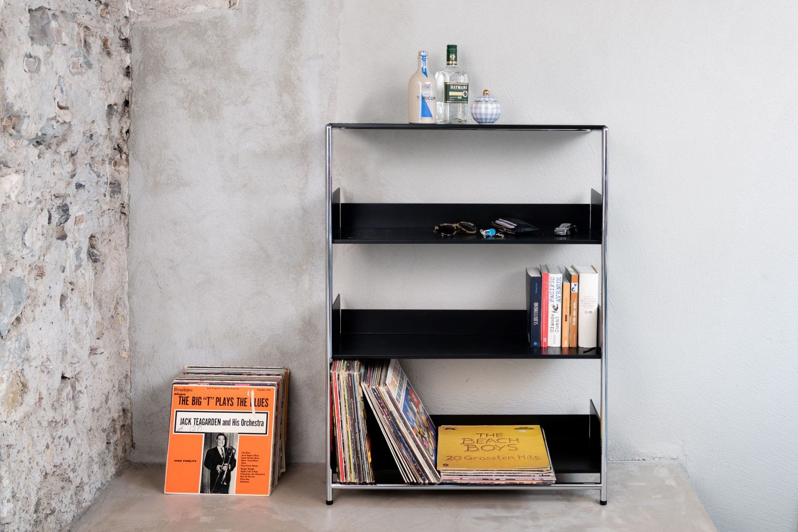 Schwarzes Pool 110 Anlehnregal von Mox mit Schallplatten, Büchern und Dekoration vor einer Betonwand.