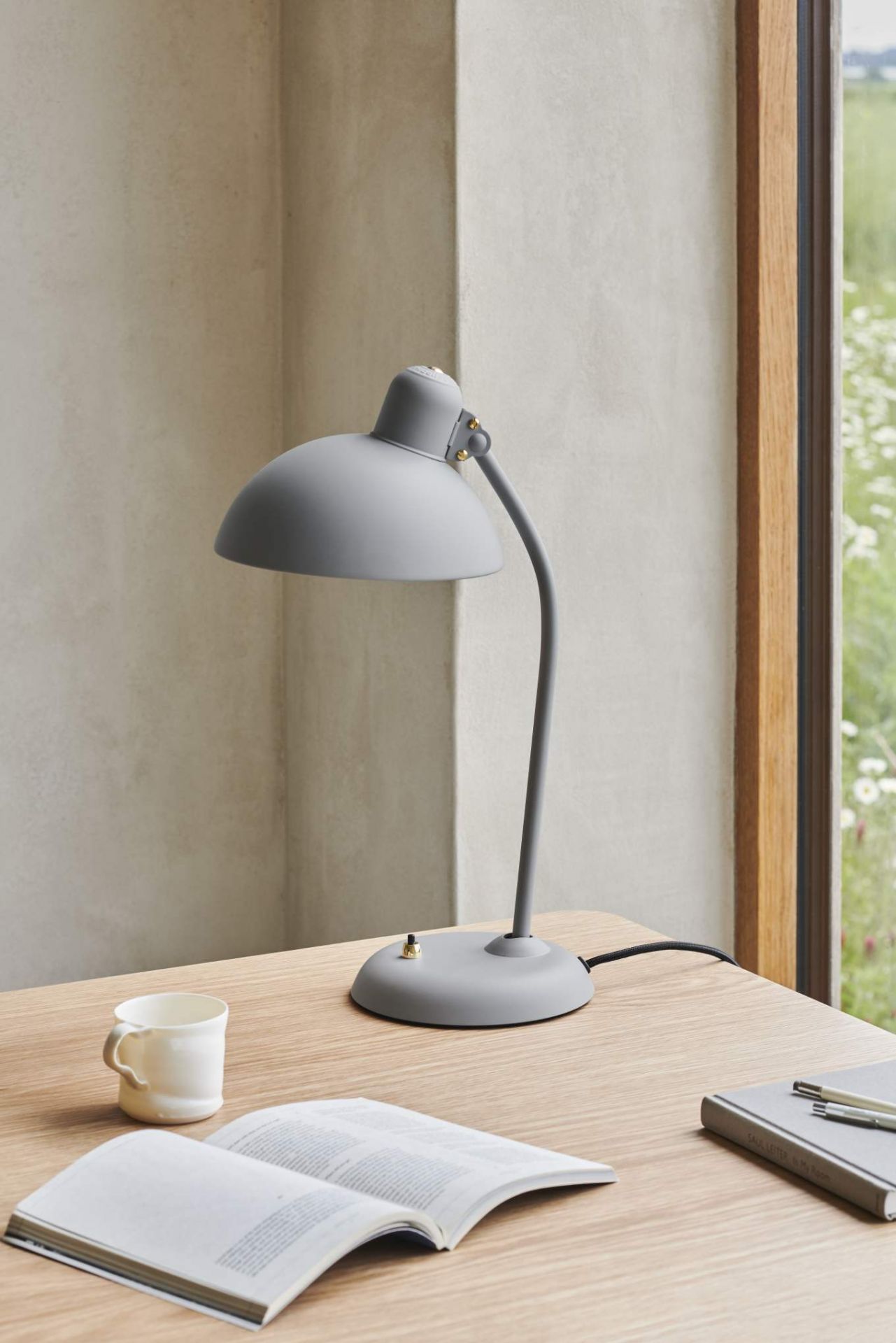 Graue KAISER idell Tischleuchte auf Schreibtisch mit Buch, Tasse und Notizbuch. Designlampe für Büro.