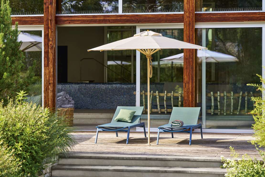 Weishäupl Klassiker Sonnenschirm über zwei blauen Liegestühlen auf einer Holzterrasse vor einem modernen Haus.