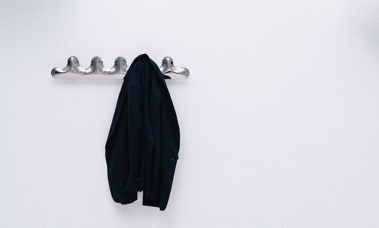 Kamm Garderobe Zieta aus poliertem Stahl an weißer Wand mit hängendem, dunklem Mantel.
