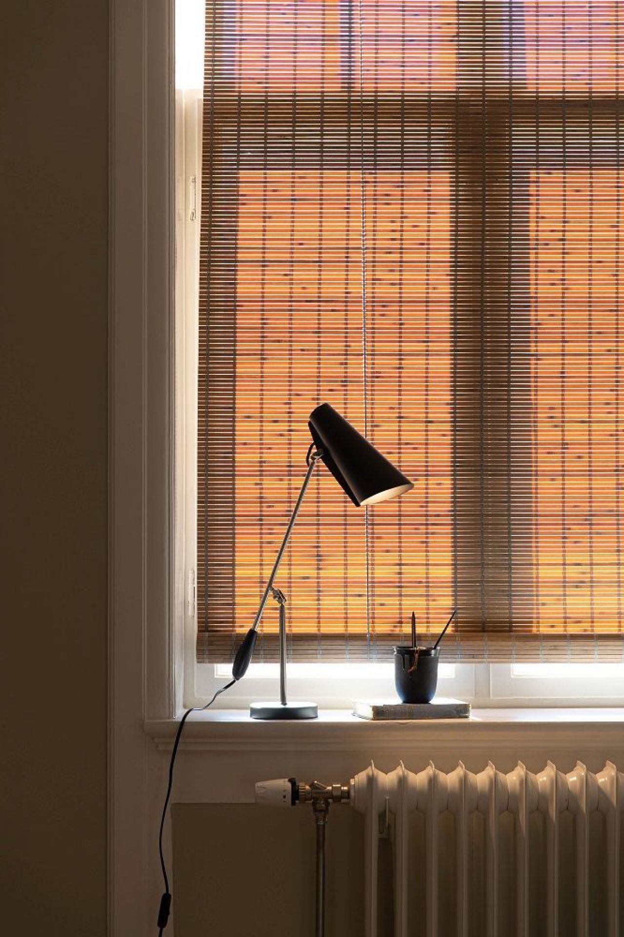 Schwarze Birdy Tischleuchte von Northern auf einer Fensterbank mit Jalousie im Hintergrund. Designlampe für Wohnzimmer.