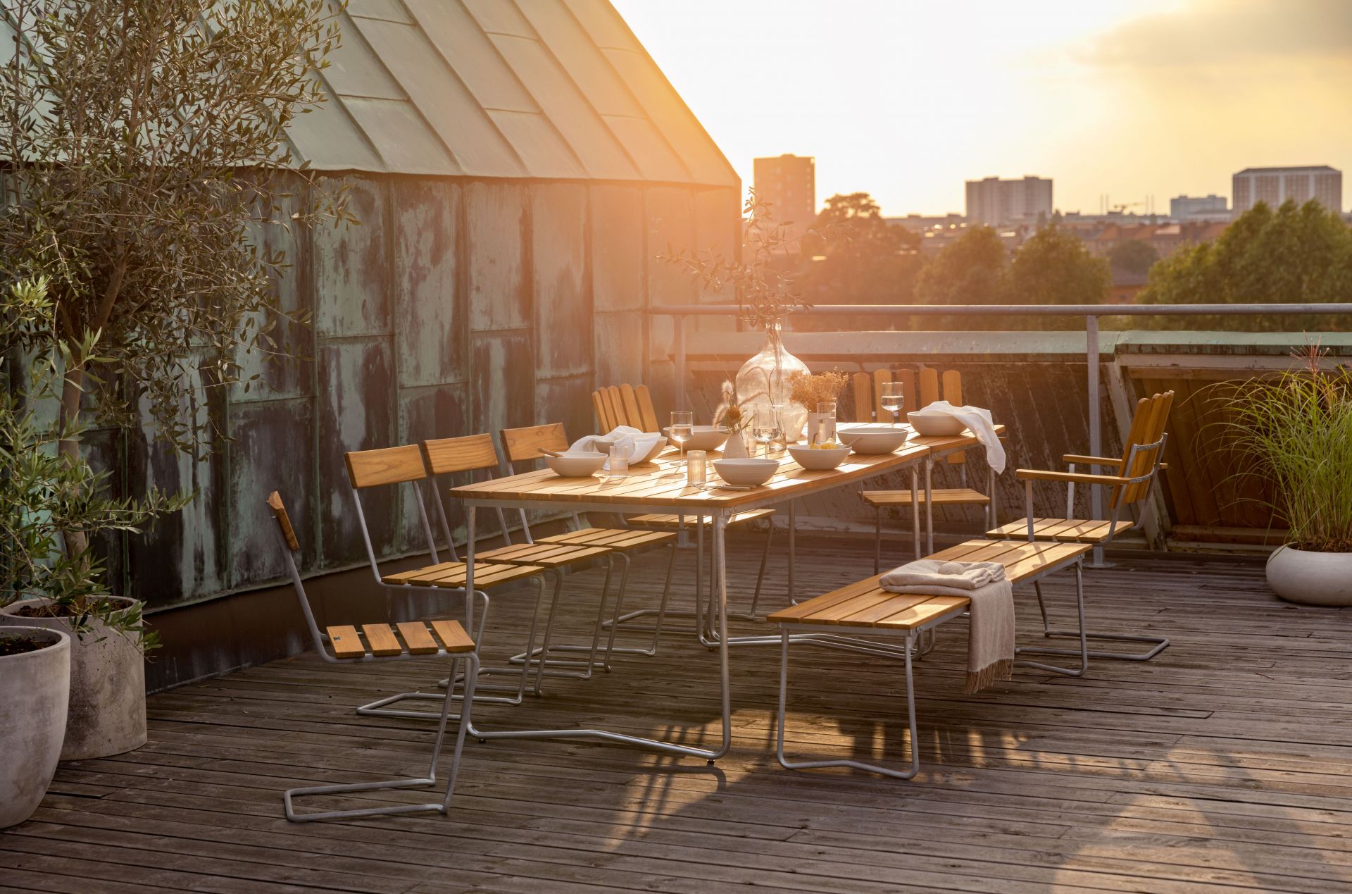 Gedeckter Esstisch mit Grythyttan Stålmöbler Bank 8 Eiche und Stühlen auf einer Dachterrasse im goldenen Abendlicht.