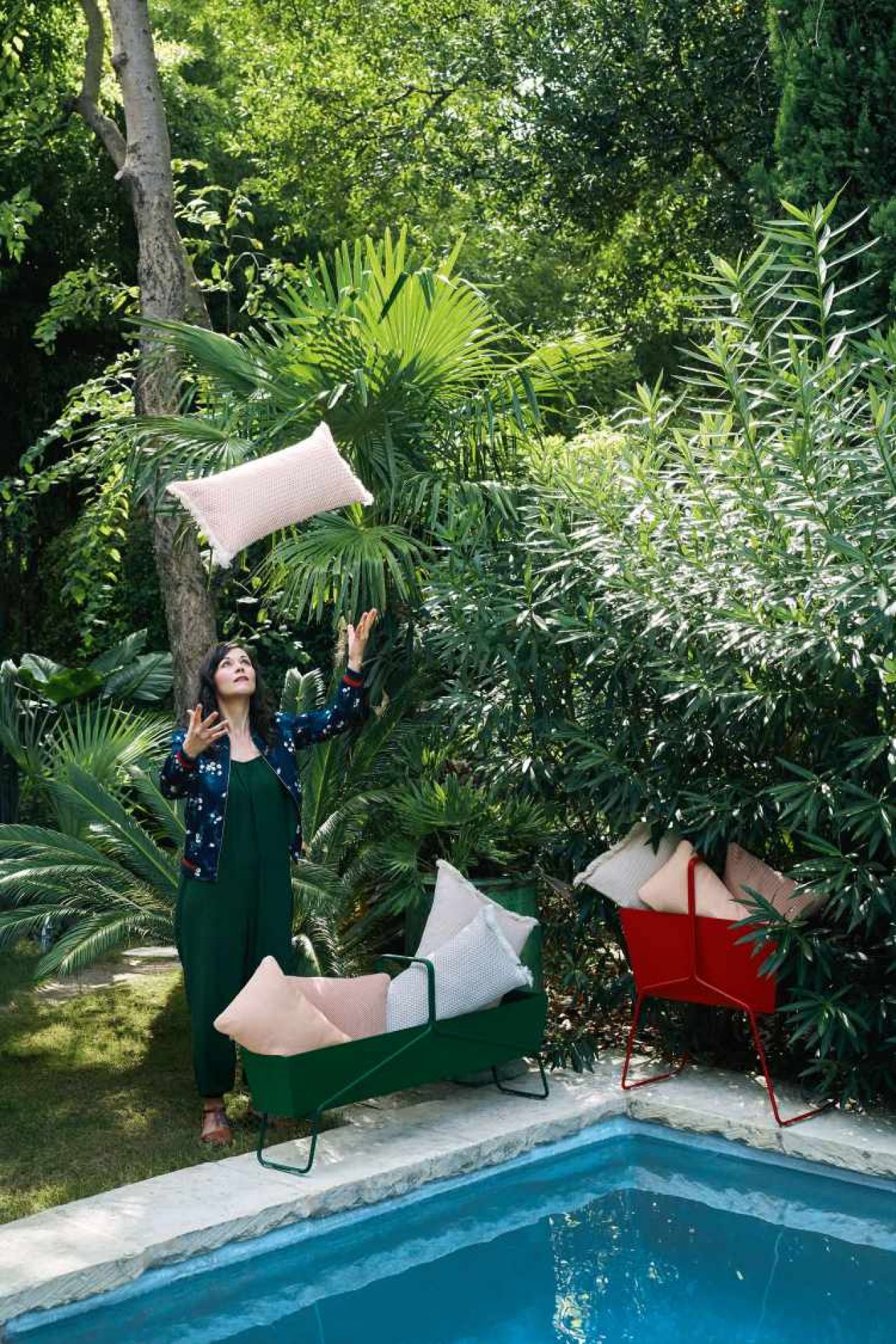 Frau wirft Kissen in Fermob Basket Blumenkasten hoch, grüne und rote Pflanzgefäße am Pool.