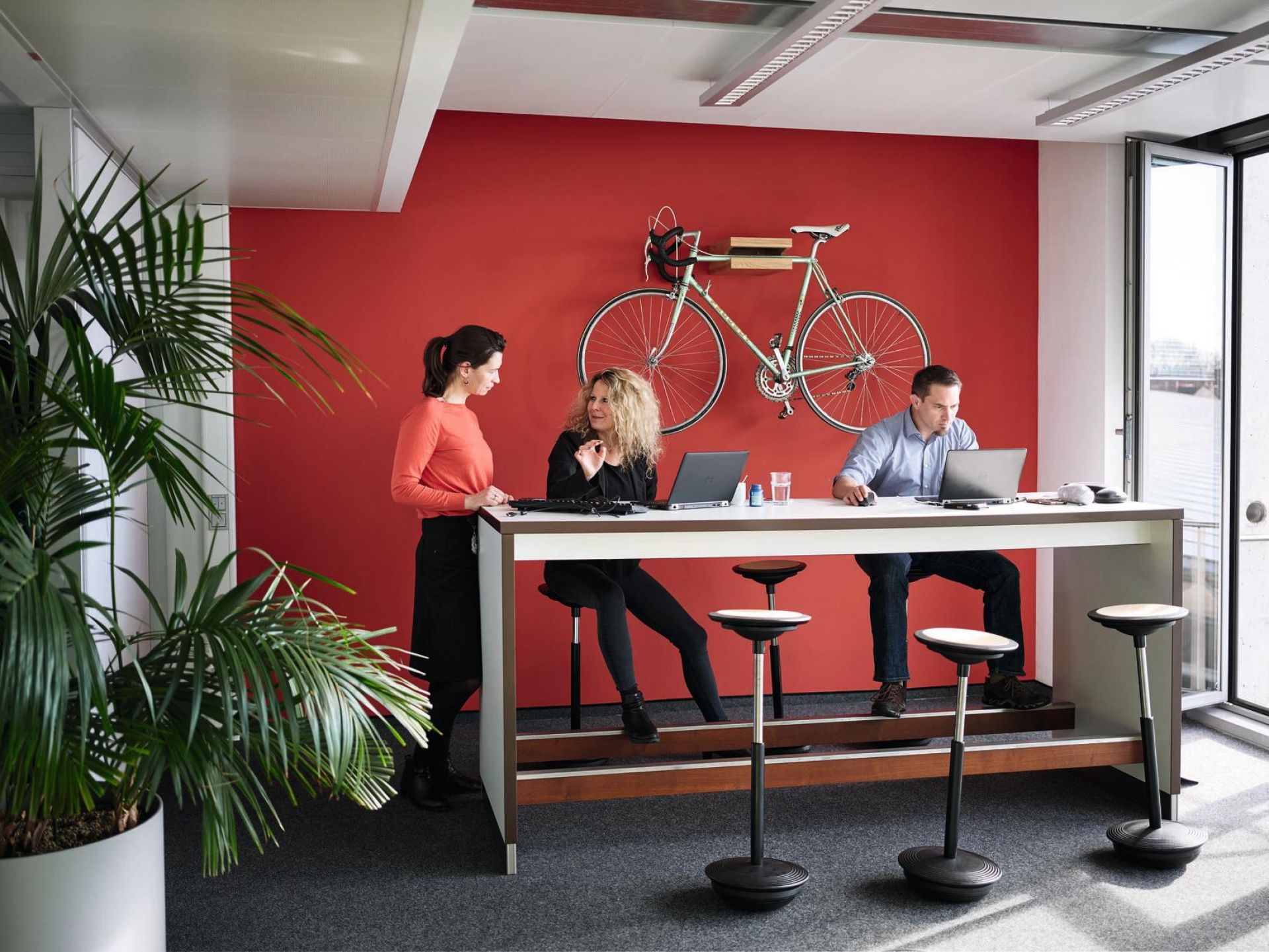 Büro mit Stehtisch und Wilkhahn Stitz Stehhockern, drei Personen arbeiten an Laptops.