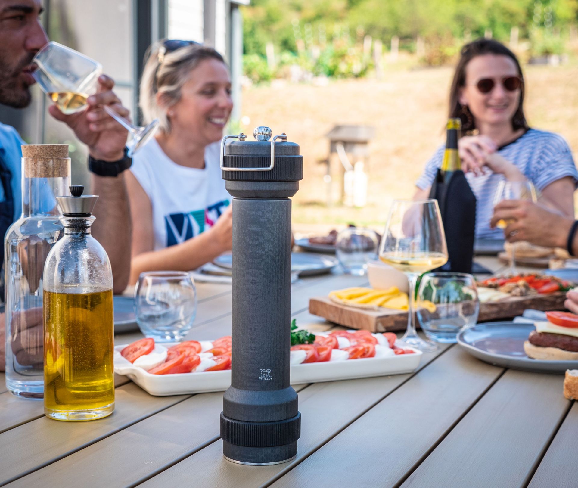 Peugeot BBQ Pfeffermühle Graphit 30 cm auf Tisch mit Speisen und Menschen im Hintergrund.