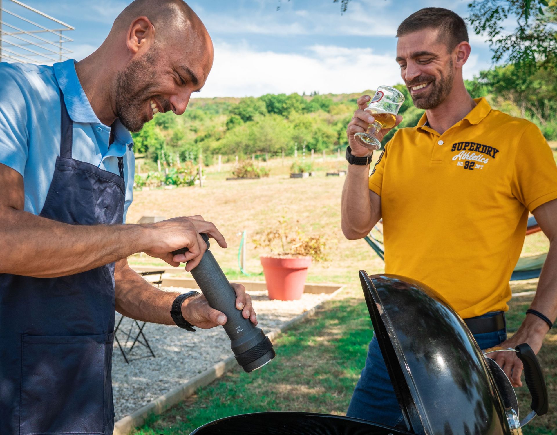 Zwei Männer grillen im Freien, einer benutzt eine Peugeot Pfeffermühle, der andere trinkt Bier.