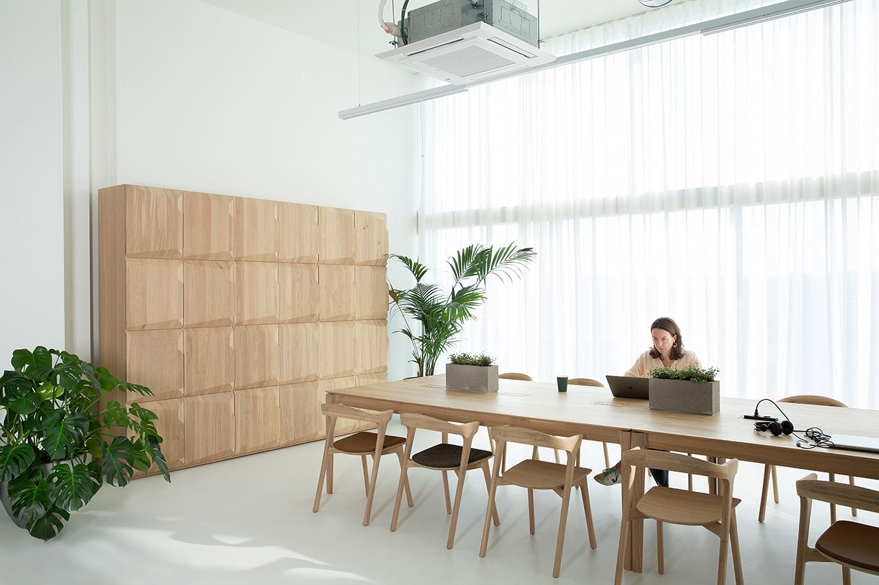 Heller Raum mit Ethnicraft Bok Cowork Schreibtisch aus Eichenholz, Stühlen und arbeitender Frau am Laptop.