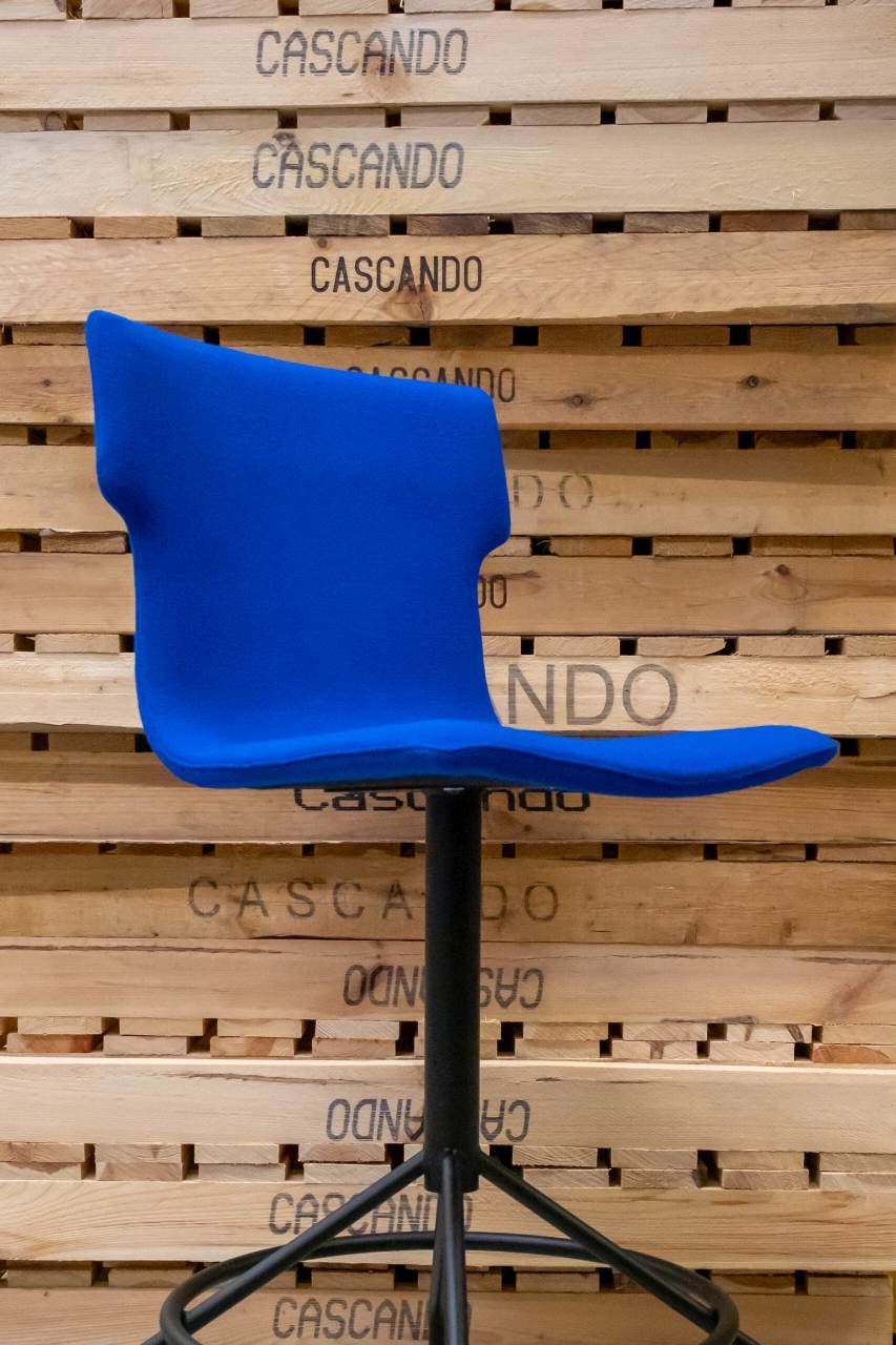 Blauer Cascando Co-Chair Bürodrehstuhl mit schwarzem Fußkreuz vor einer Holzwand mit Firmenlogo.