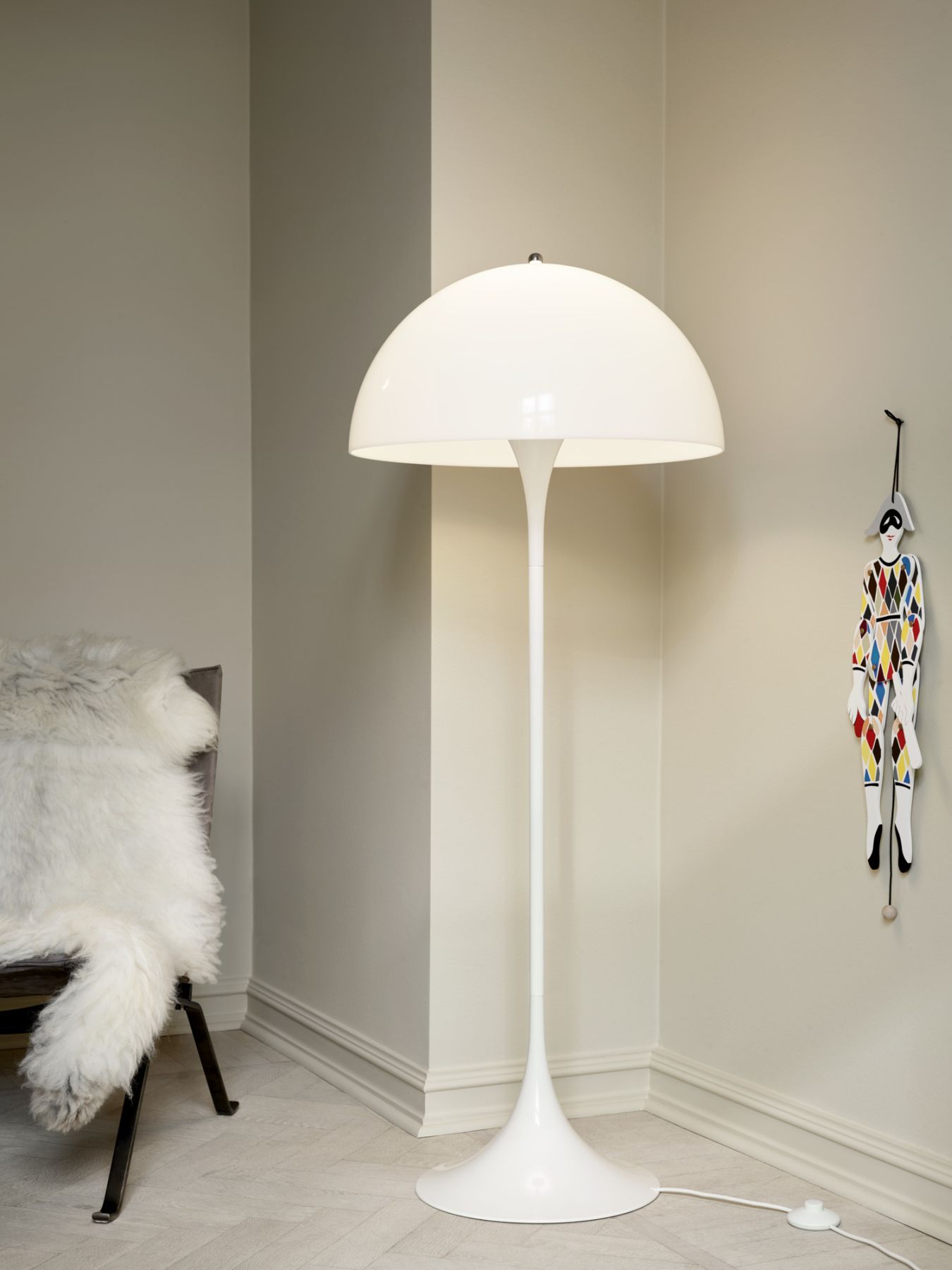 Moderne, weiße Stehlampe im Pilzdesign neben einem Sessel mit Fell und einer Harlekin-Figur an der Wand.