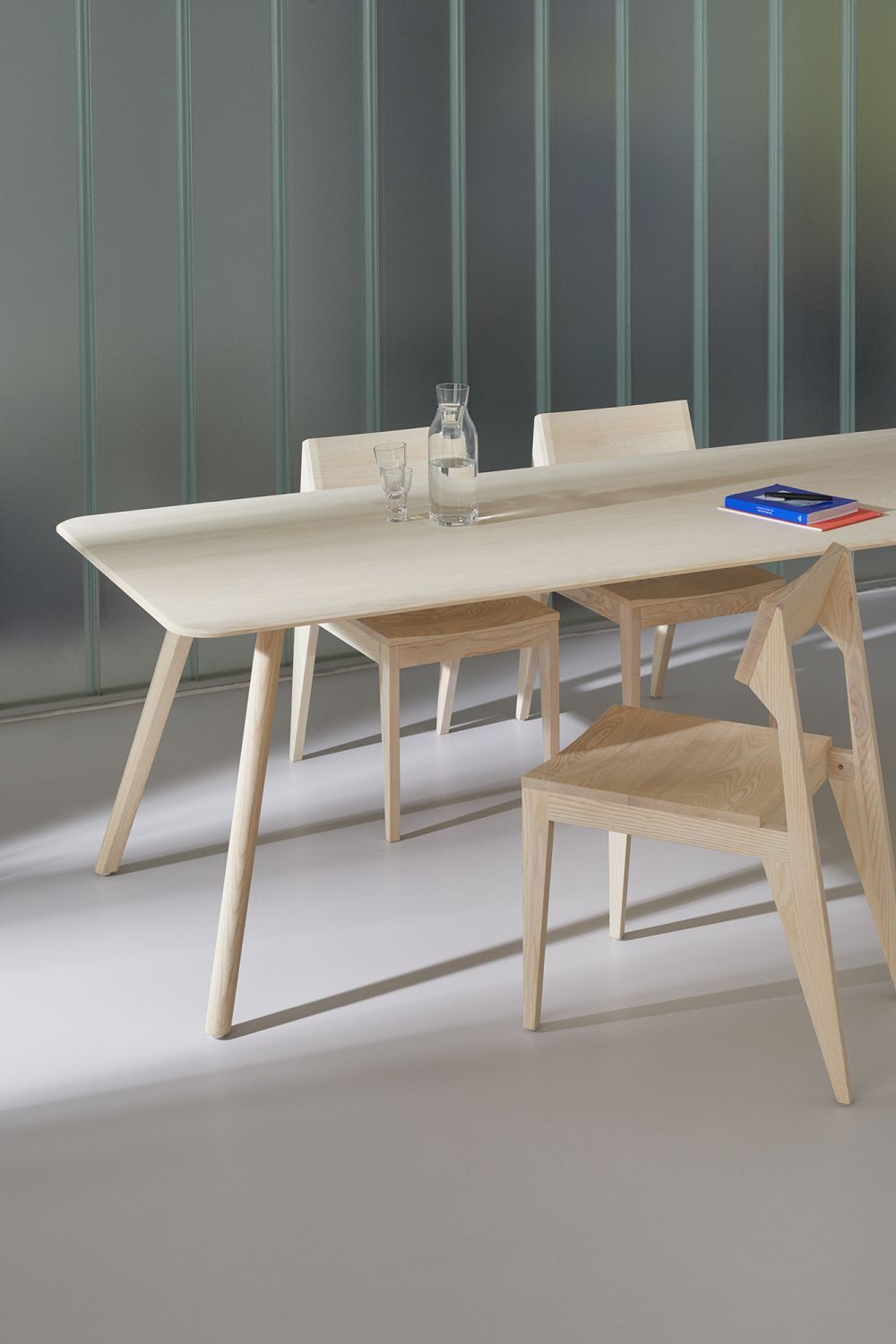 MEYER Tisch Large OUT aus hellem Holz mit Stühlen, Wasserflasche und Buch auf dem Tisch.