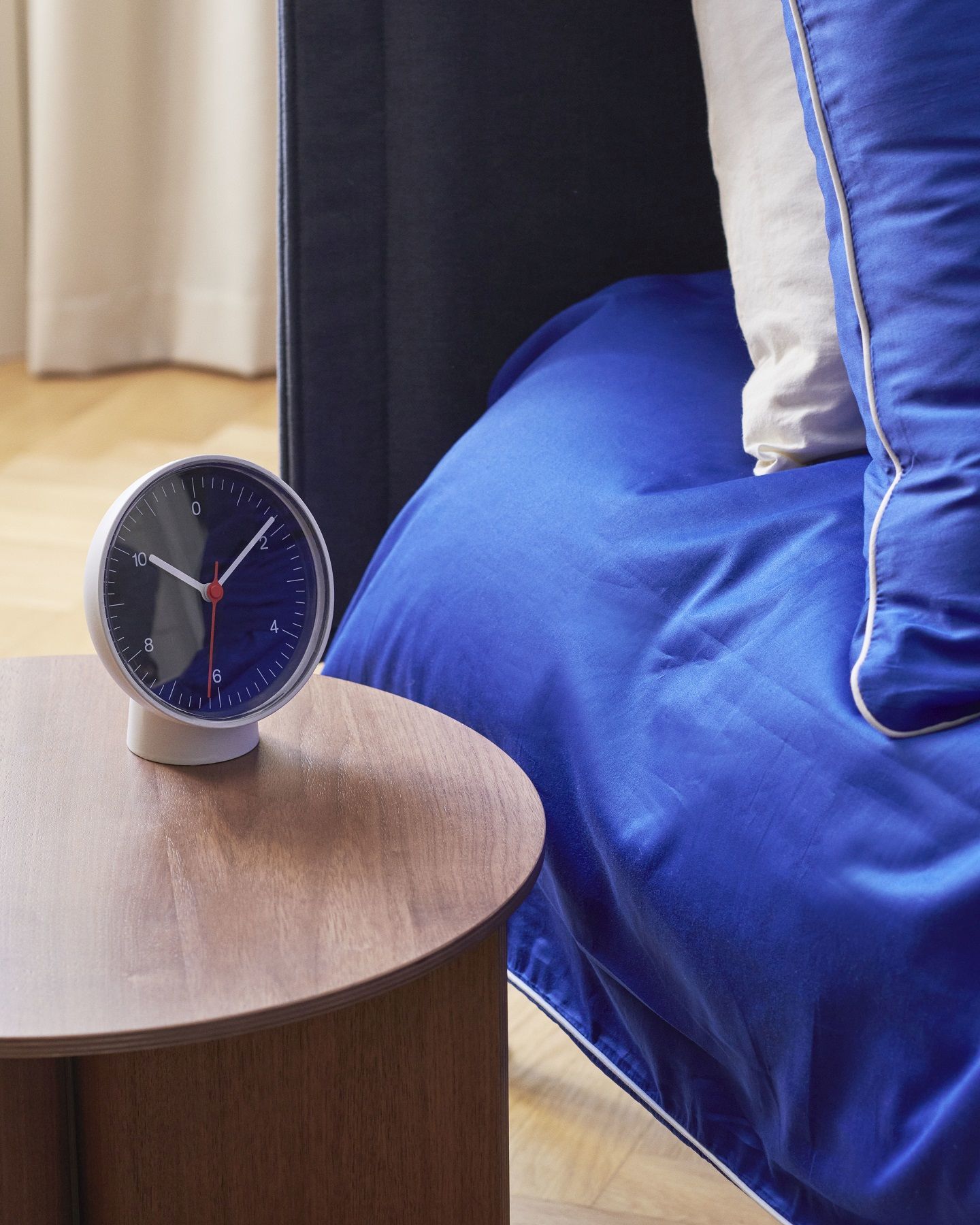 Hay Tischuhr Dual-function auf einem Nachttisch neben einem Bett mit blauer Bettwäsche.