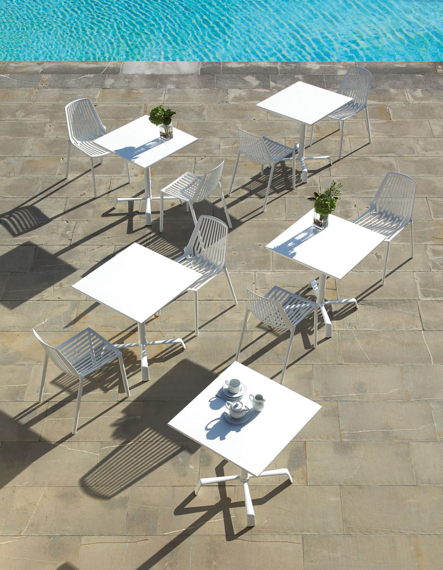 Mehrere weiße Rion Outdoor Stühle und Tische auf einer Terrasse neben einem Pool. Gartenmöbel-Set.