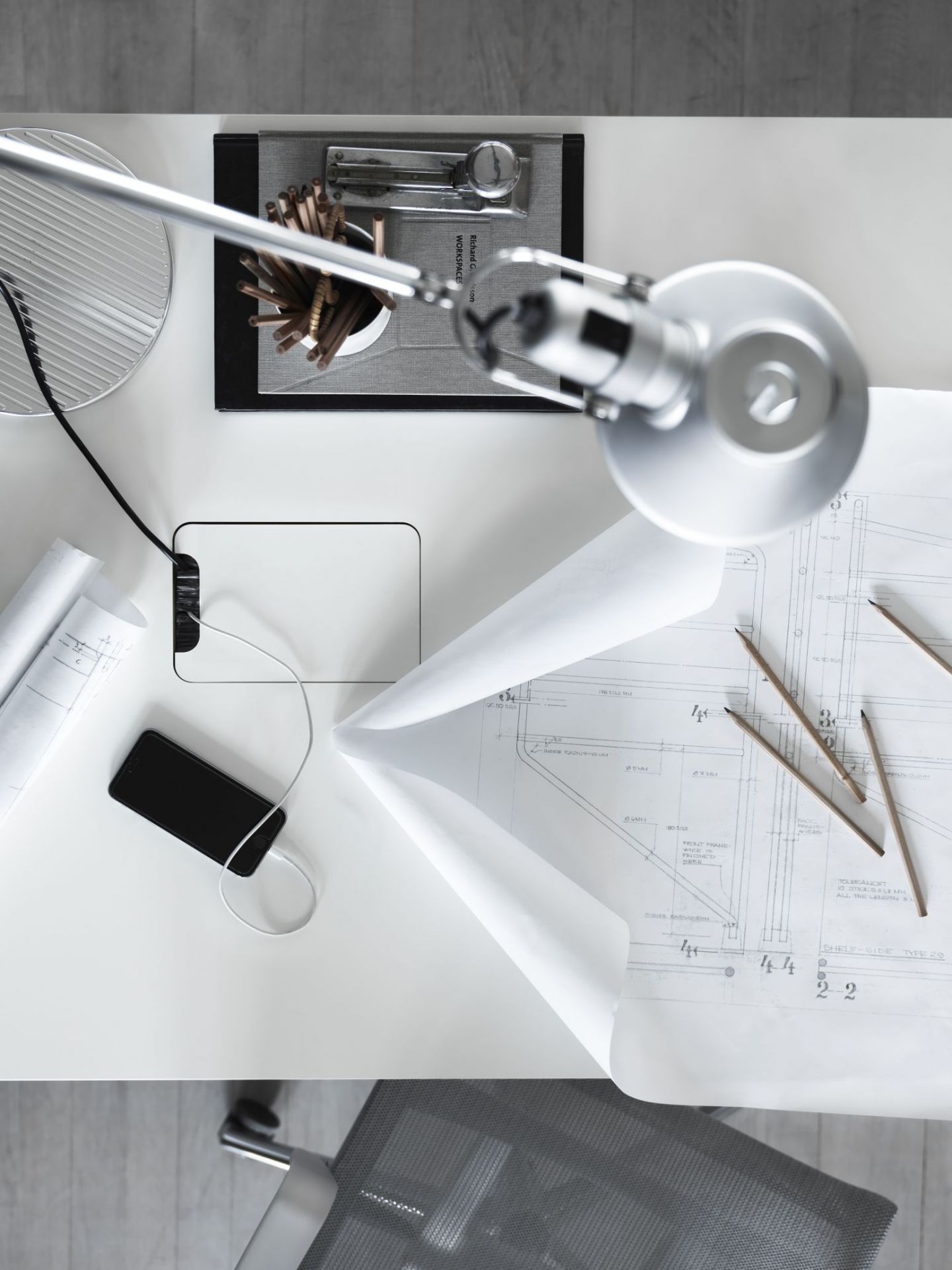 Draufsicht auf einen weißen Schreibtisch von String Furniture mit Lampe, Handy und Bauplänen.