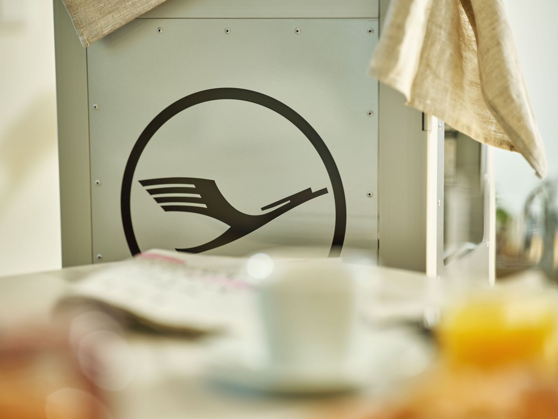 Lufthansa Bordbar Trolley mit Logo, Handtuch, Kaffeetasse und Orangensaft im unscharfen Vordergrund.