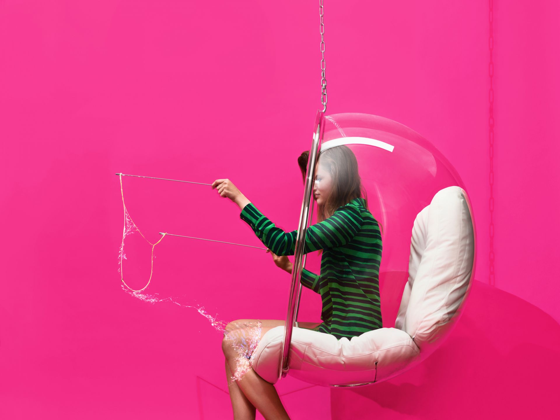 Frau sitzt im transparenten Bubble Chair Hängesessel von Eero Aarnio und macht Seifenblasen vor pinkem Hintergrund.