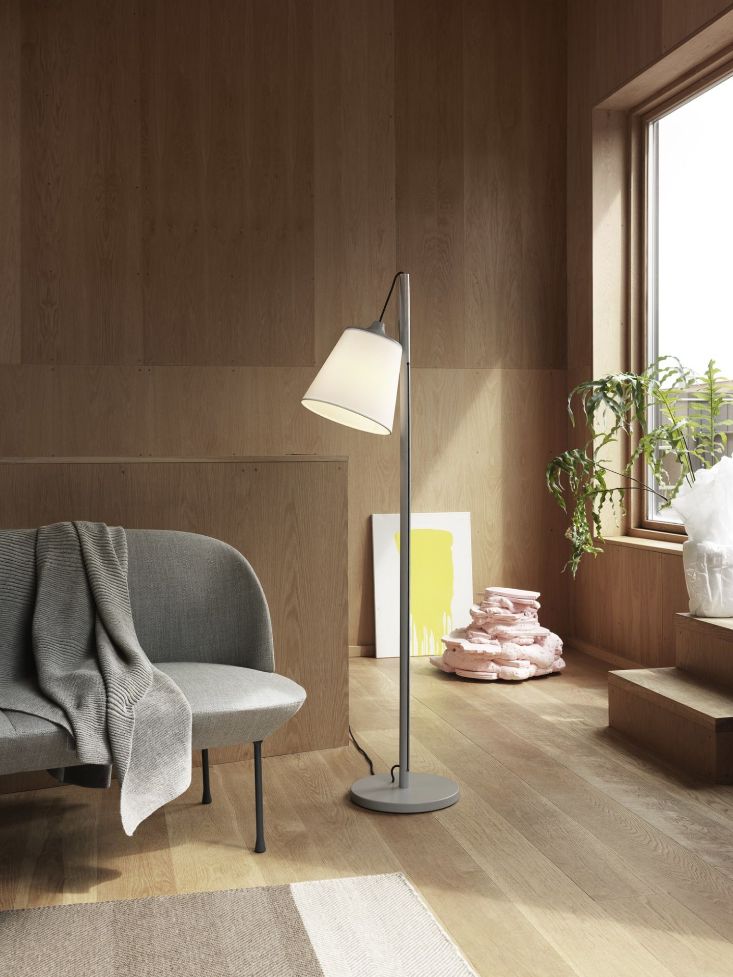 Heller Wohnraum mit Oslo Sofa, Stehlampe und Kunstobjekten vor einer Holzvertäfelung und Fenster.