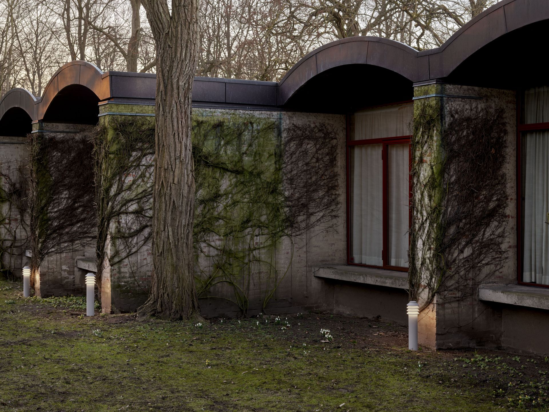 Bysted Garden Pollerleuchte von Louis Poulsen im Außenbereich, stilvolle Gartenbeleuchtung neben einem Gebäude mit Bewuchs.