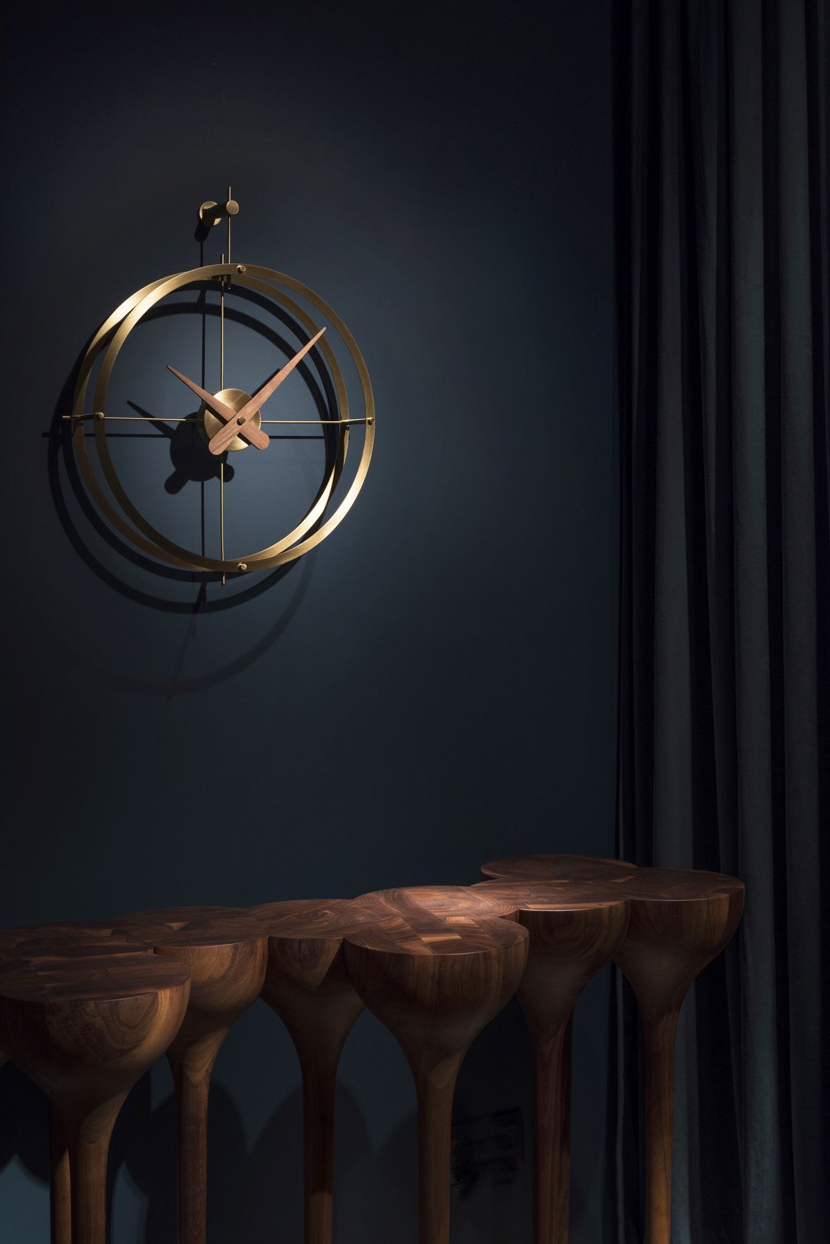 Nomon 2 Puntos Gold Uhr, moderne Wanduhr mit goldenem Rahmen und Holzzeigern vor dunkler Wand.