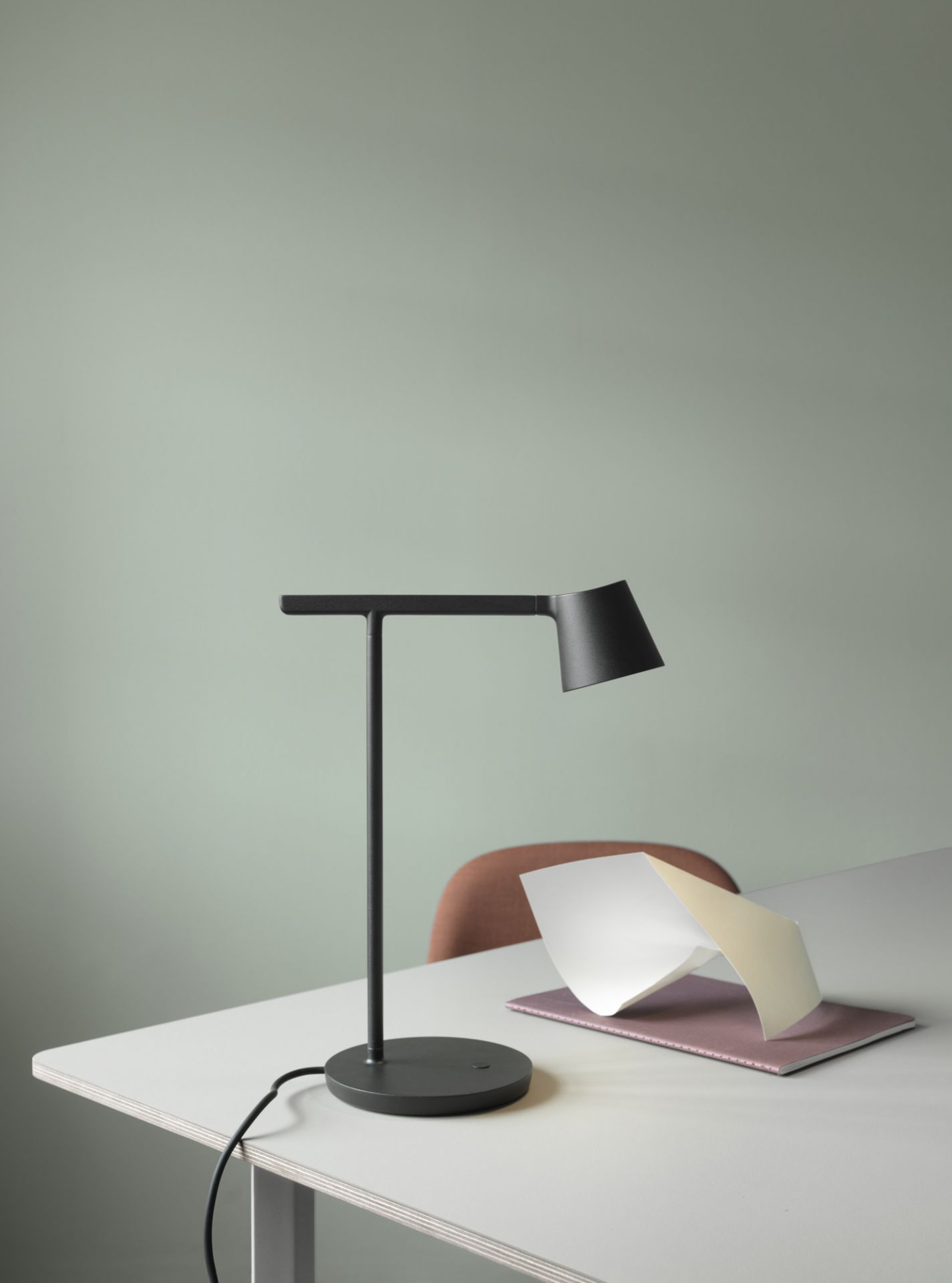 Schwarze Tip Table Lamp Tischleuchte von Muuto auf einem Schreibtisch mit Notizbuch vor hellgrünem Hintergrund.