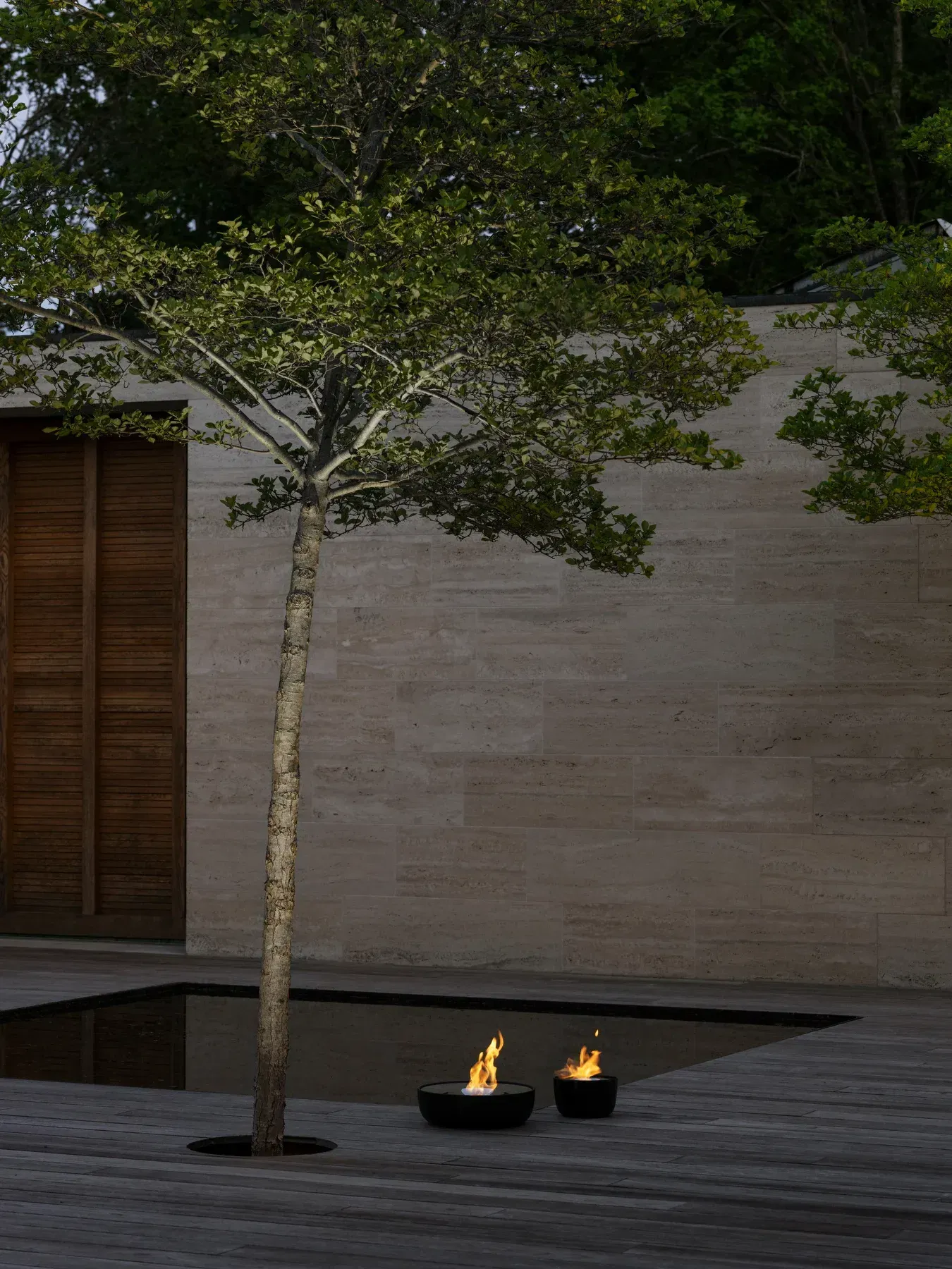 Zwei schwarze Feuerstellen auf Holzterrasse mit Baum und Wasserbecken für stimmungsvolle Gartenbeleuchtung.
