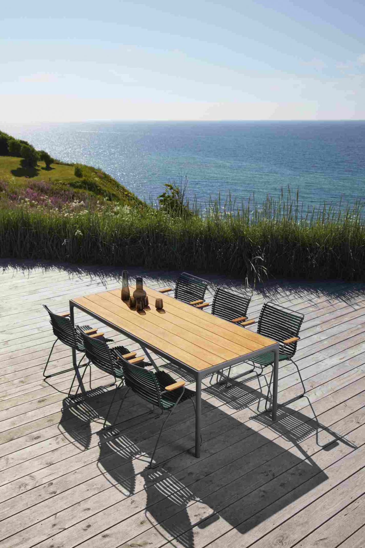 Dunkelgraue Click Dining Stühle mit Armlehne von Houe und Esstisch auf Holzterrasse mit Meerblick.