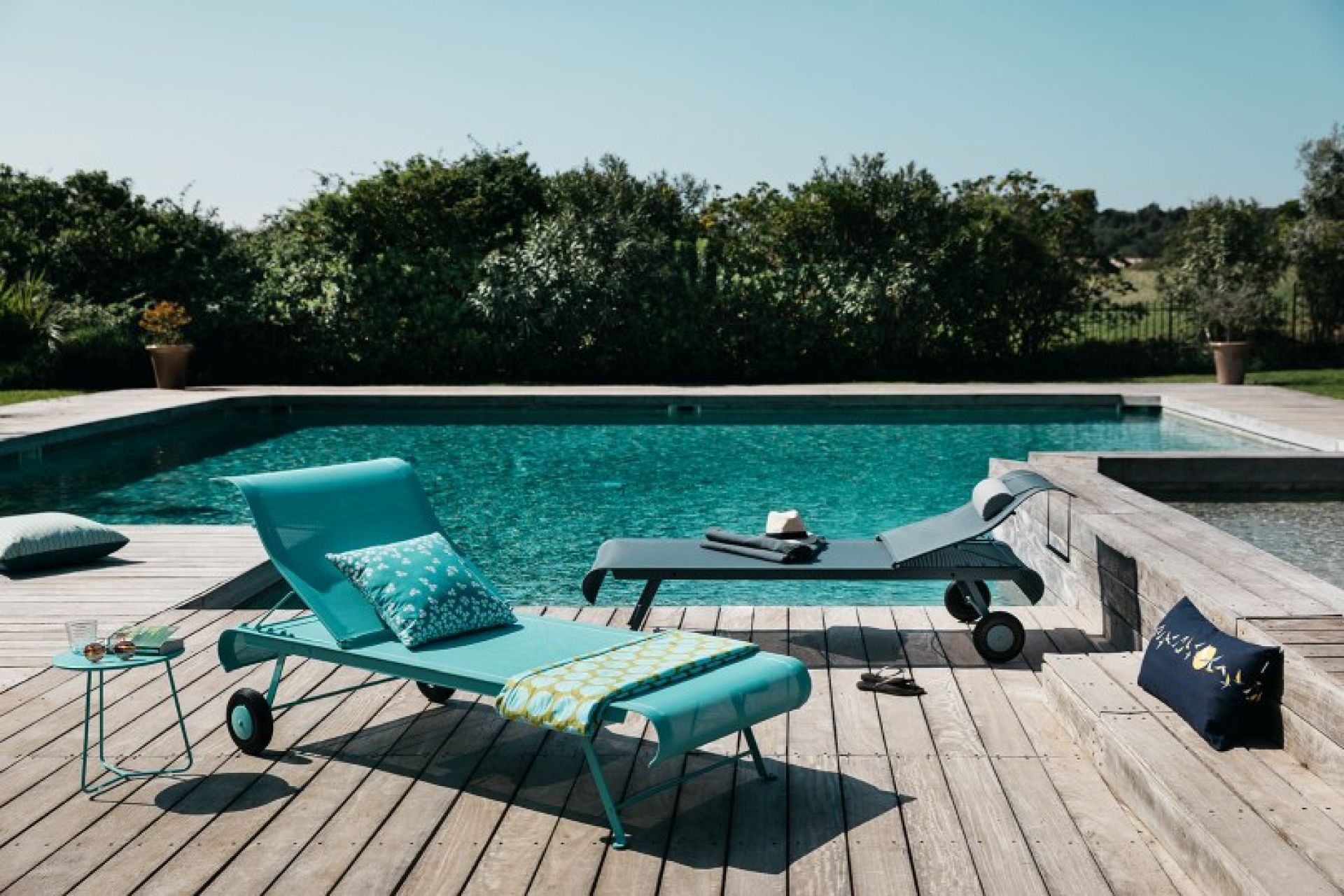 Türkisblaue Dune Sonnenliege von Fermob am Pool auf einer Holzterrasse mit Kissen.