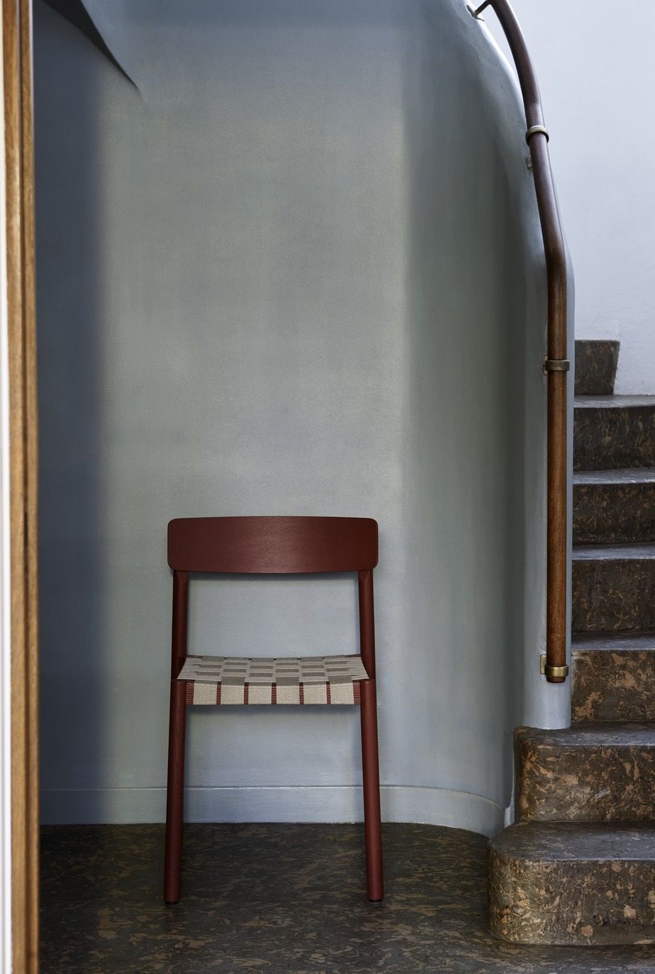 AndTradition Betty TK1 Stuhl in Burgunderrot mit gewebter Sitzfläche vor Treppenhaus. Designstuhl für modernes Wohnen.