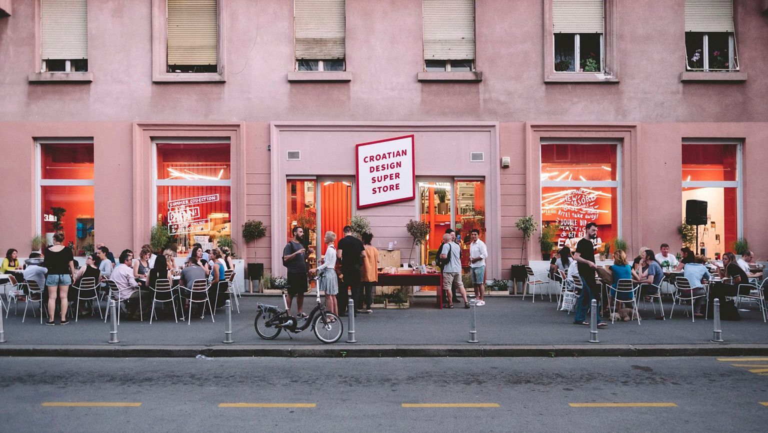 Außenansicht eines Cafés mit Prostoria Stühlen, Tischen und Gästen vor einem rosa Gebäude.