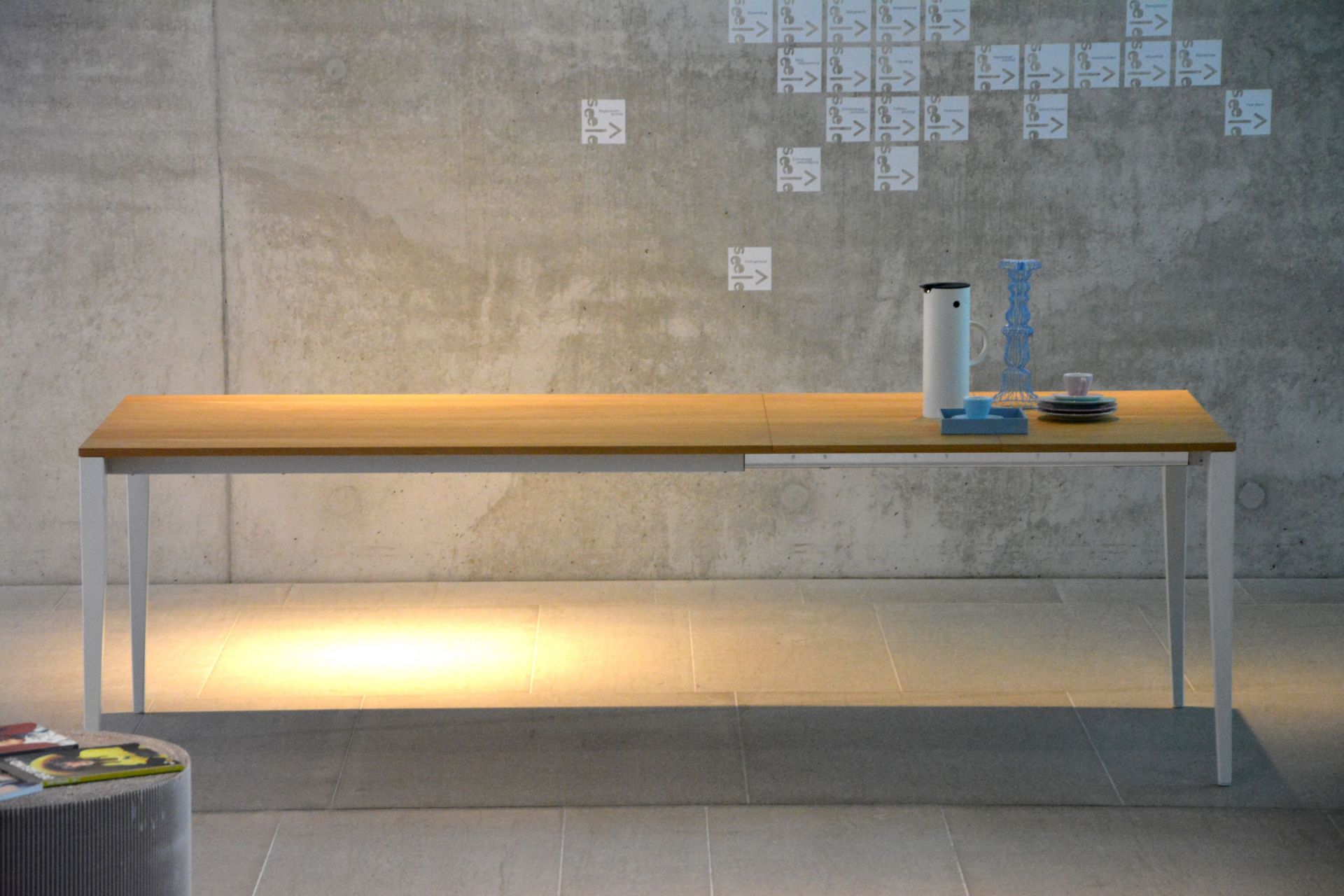 Jupiter Ausziehtisch von Jan Kurtz mit heller Holzplatte und weißem Metallgestell vor Betonwand.
