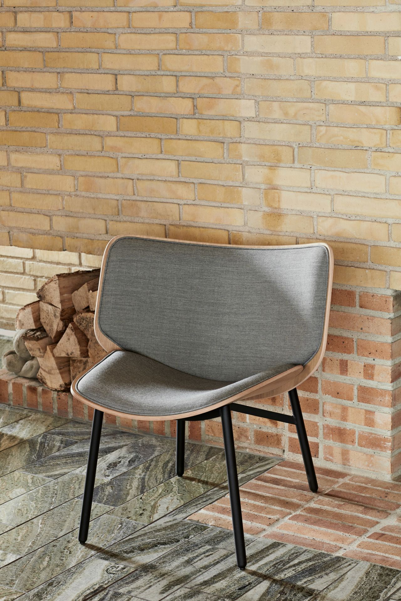 Grauer Dapper Lounge Chair Sessel von Hay mit Holzrahmen und schwarzen Beinen vor Ziegelwand.