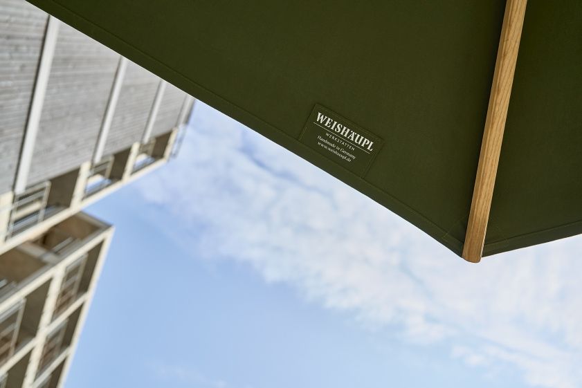 Detailaufnahme des Weishäupl Klassiker Sonnenschirms in Grün mit Holzstreben und Blick auf ein Gebäude.