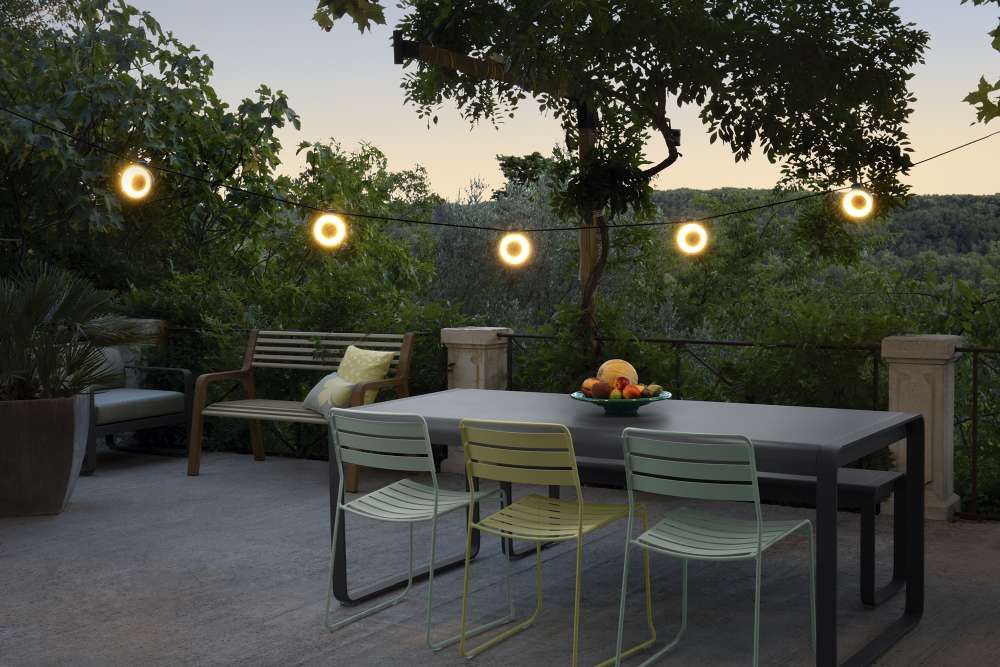 Somerset Bank Outdoor Fermob: Terrasse mit Gartenmöbeln, Tisch, Stühlen, Bank und Lichterkette im Grünen.