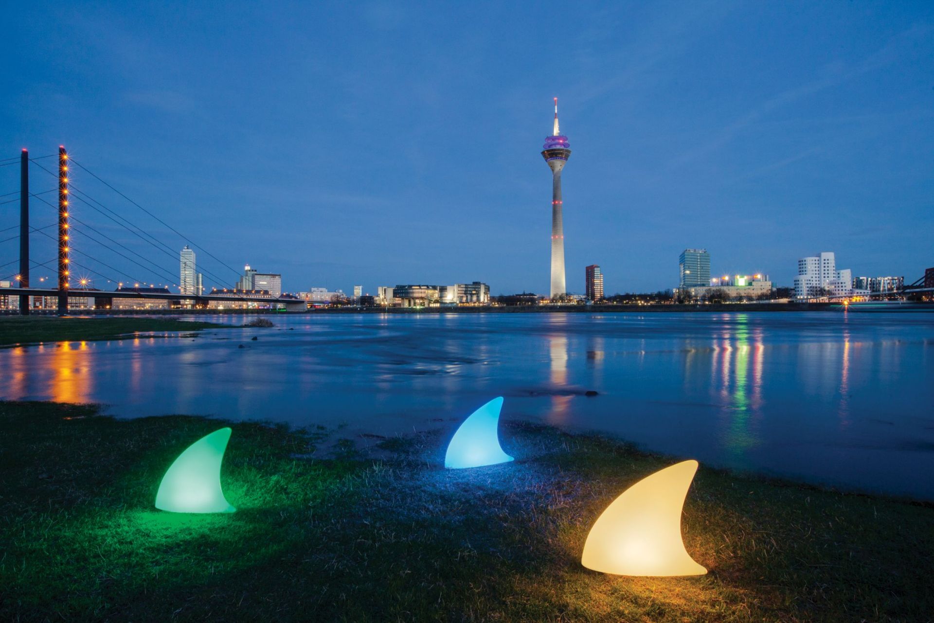 Moree Shark Haifischflosse Bodenleuchte, drei farbige LED-Leuchten am Flussufer mit Skyline im Hintergrund.