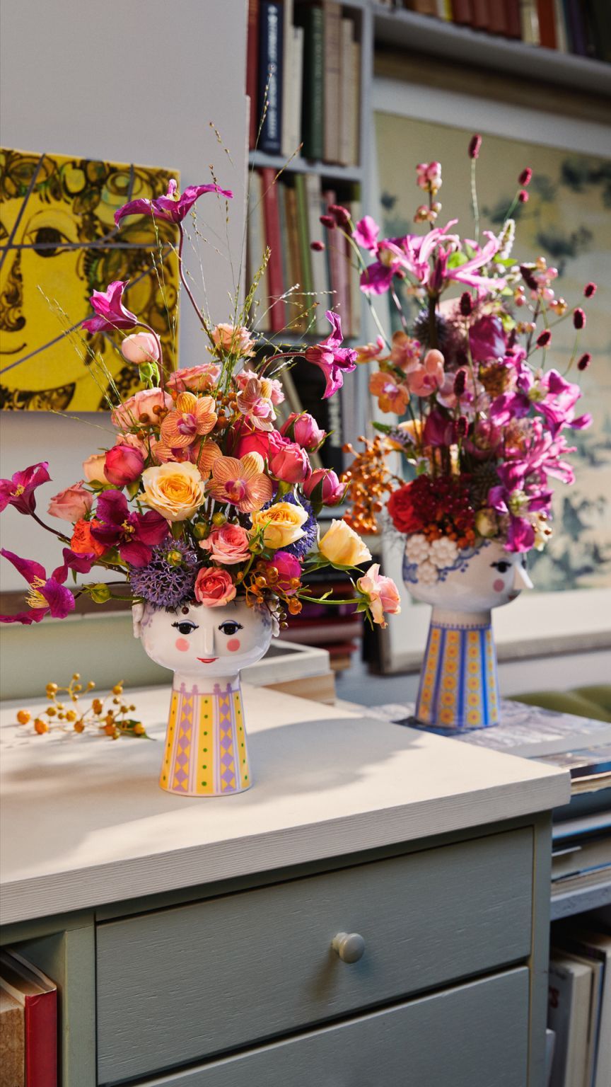 Zwei hellblaue Bjørn Wiinblad Eva Vasen (H18,5cm) mit bunten Blumenarrangements auf einem Tisch.