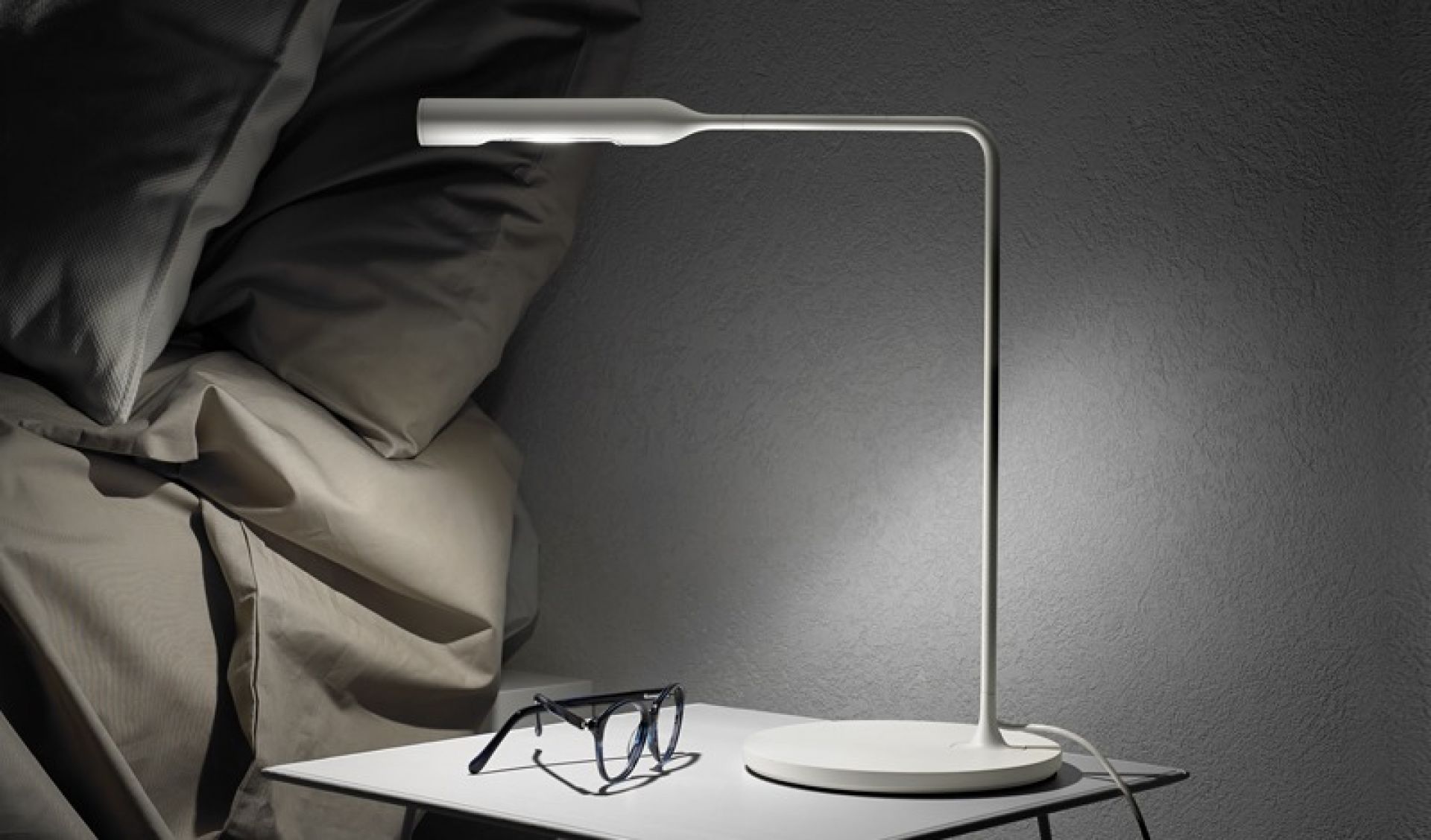 Weiße Flo Nachttischleuchte Lumina auf Nachttisch mit Brille, modernes Schlafzimmer-Ambiente.