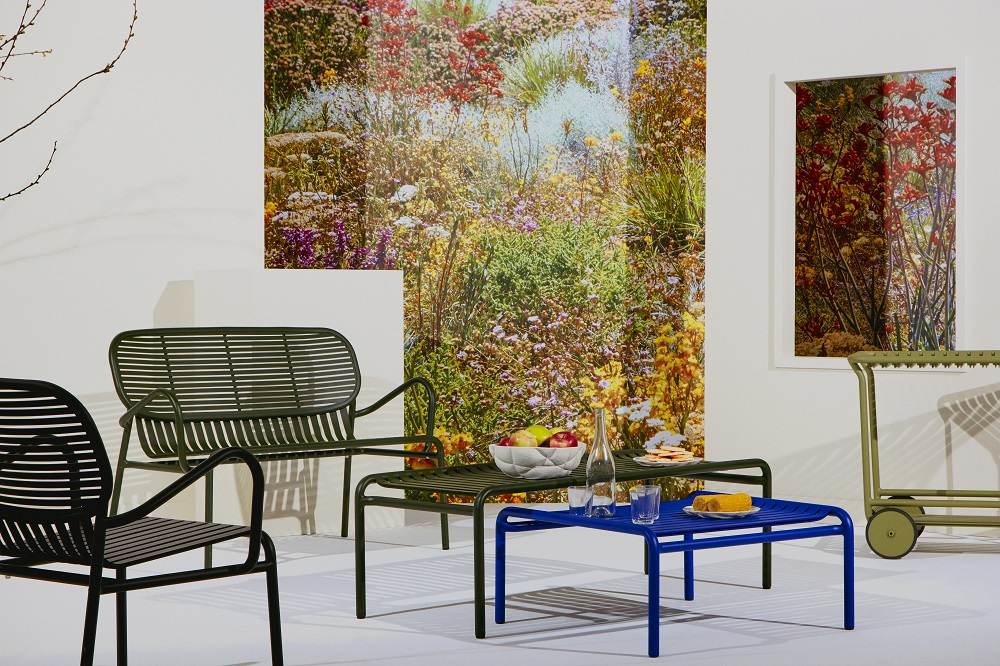 Blaues Petite Friture Week-End Sofa 2-Sitzer und Gartentisch mit Obst und Getränken vor Blumenwand.