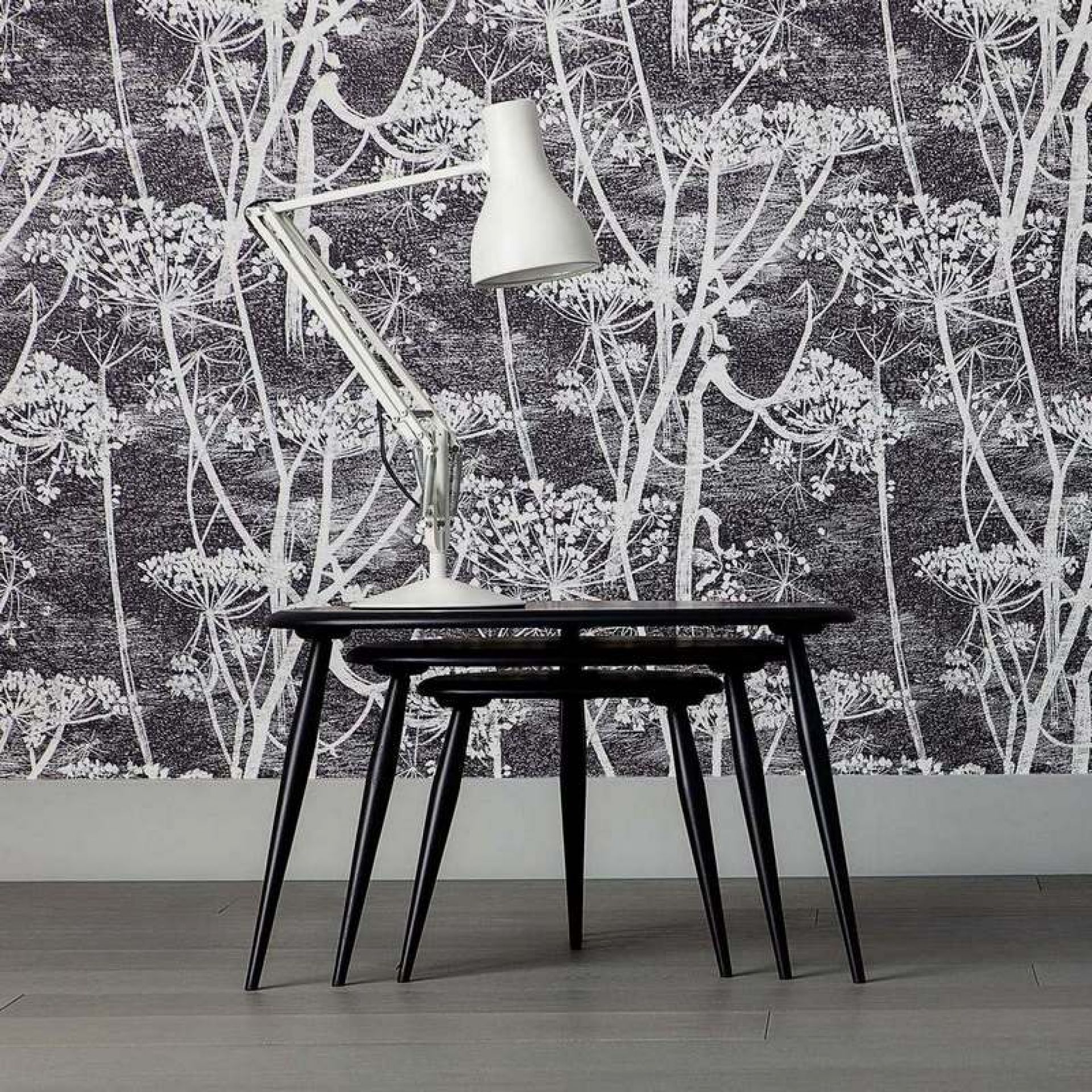 Weiße Anglepoise Type 75 Tischleuchte auf schwarzem Satztisch vor floral gemusterter Tapete.
