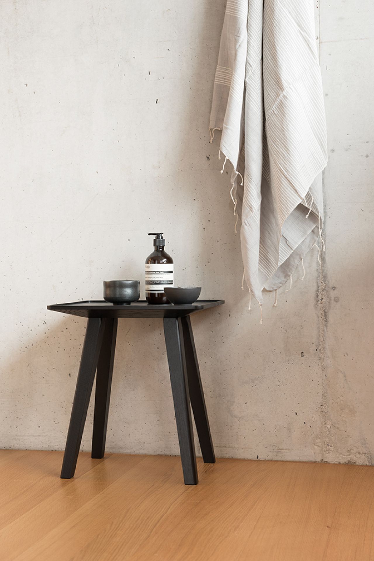 Schönbuch Nini Hocker in Schwarz mit Seife, Schalen und Handtuch vor einer Betonwand.