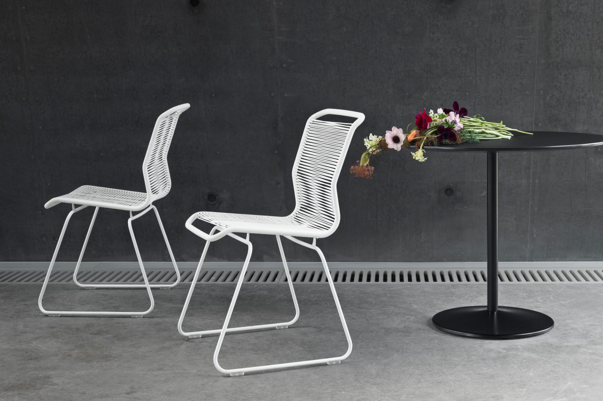 Zwei weiße Stühle und ein runder, schwarzer Montana Tisch mit Blumenstrauß vor grauer Wand.