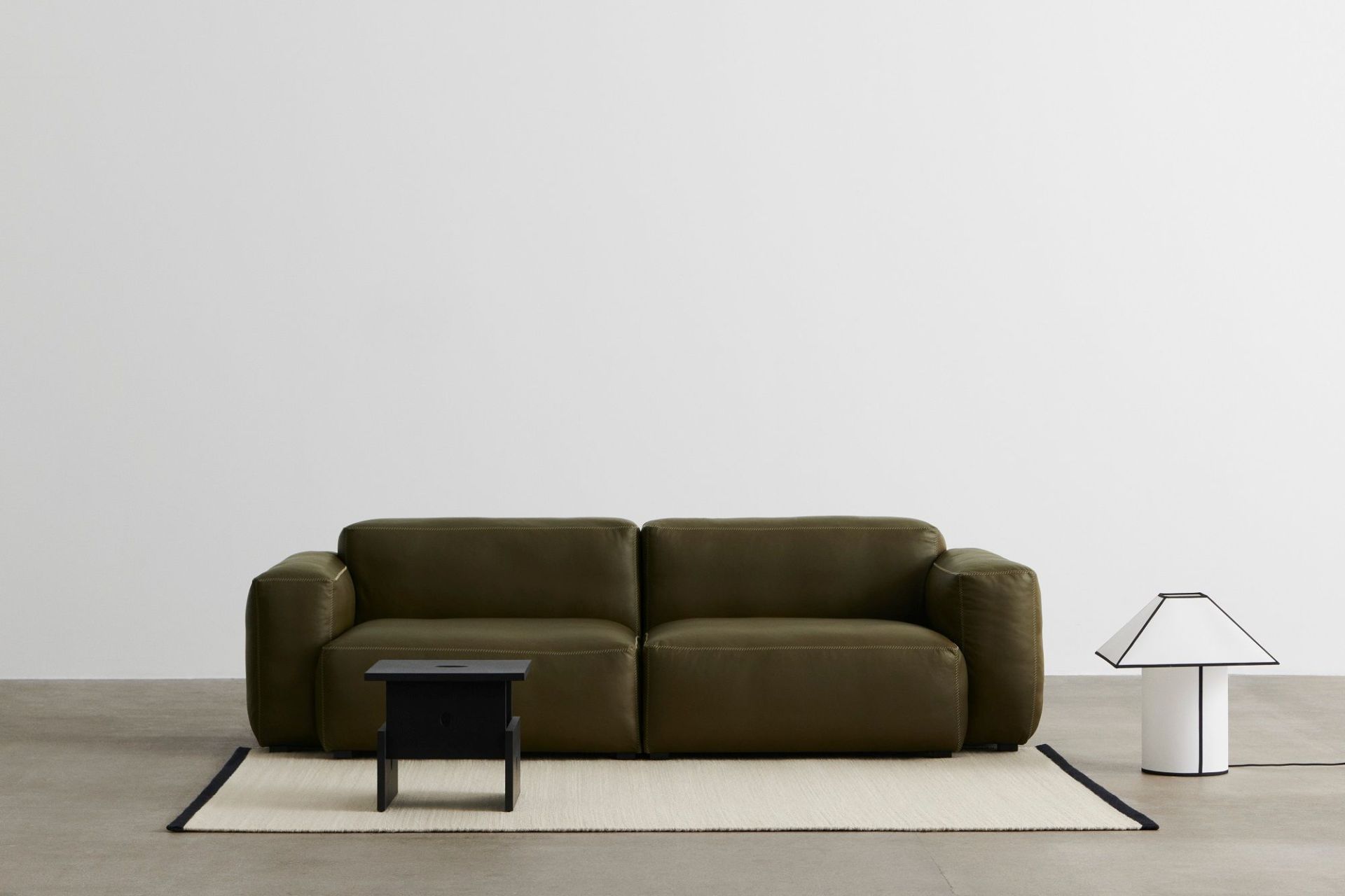 Ava Pyramid Tischleuchte von Hay, weiße Lampe mit schwarzem Rand neben olivgrünem Sofa.