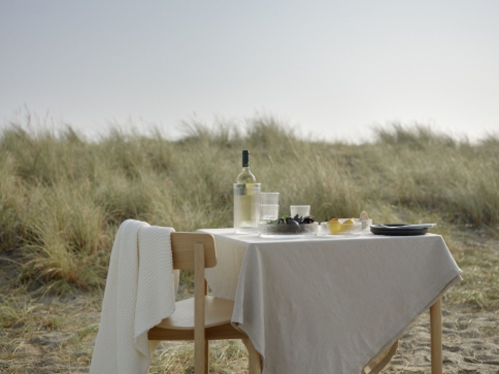 Esstisch am Strand mit Stelton Pilastro Servierschale, Wein, Gläsern und Speisen. Dekorative Tischdeko.