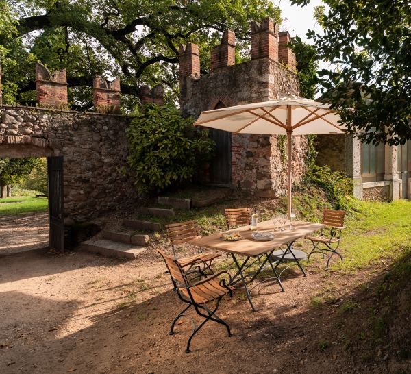 Weishäupl Klassiker Sonnenschirm über einem Gartentisch mit Stühlen im Freien vor einer alten Burgmauer.