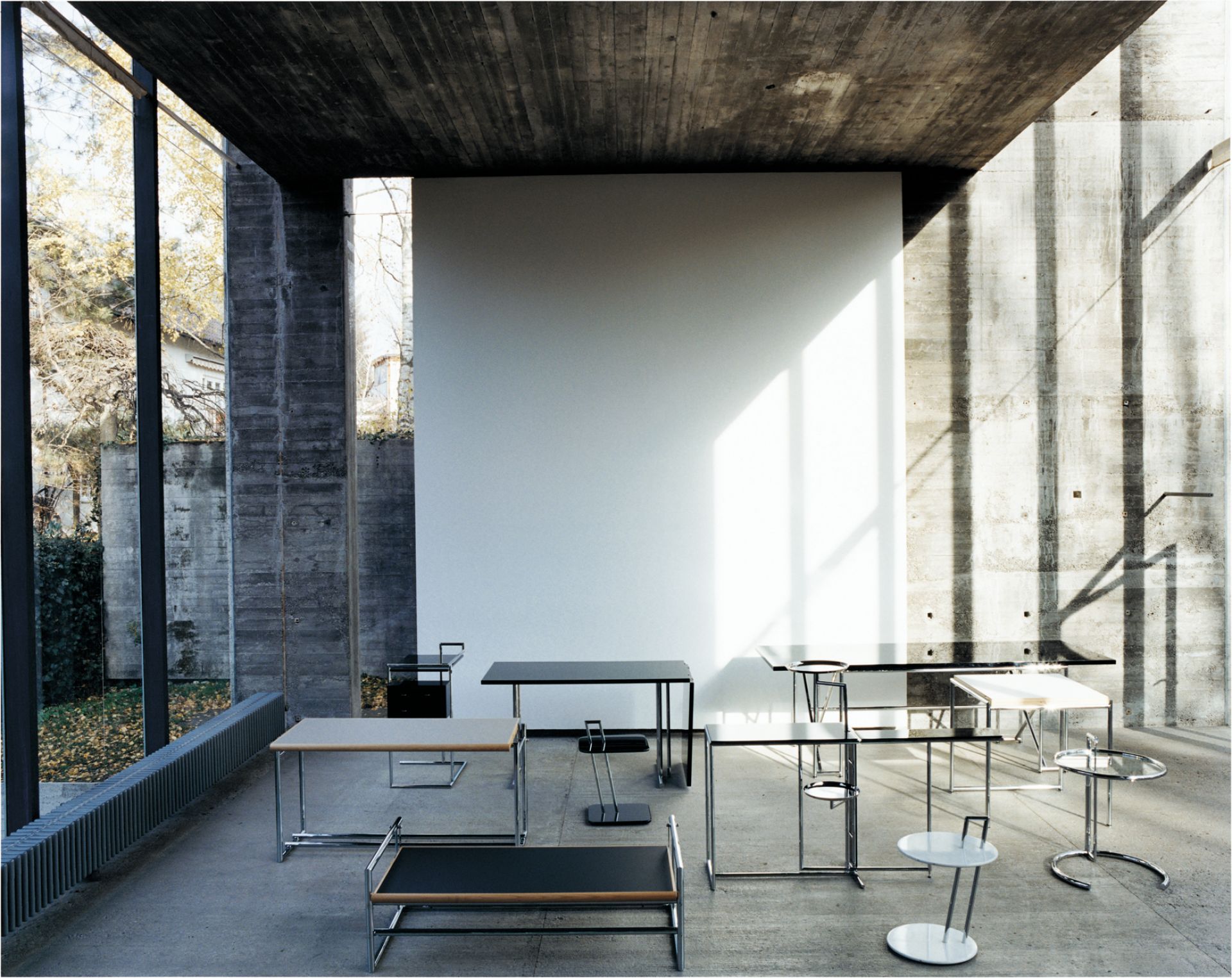 Verschiedene ClassiCon Tische, darunter der E1027 Beistelltisch, in einem modernen Raum mit Betonwänden. Designmöbel für Wohnen und Büro.