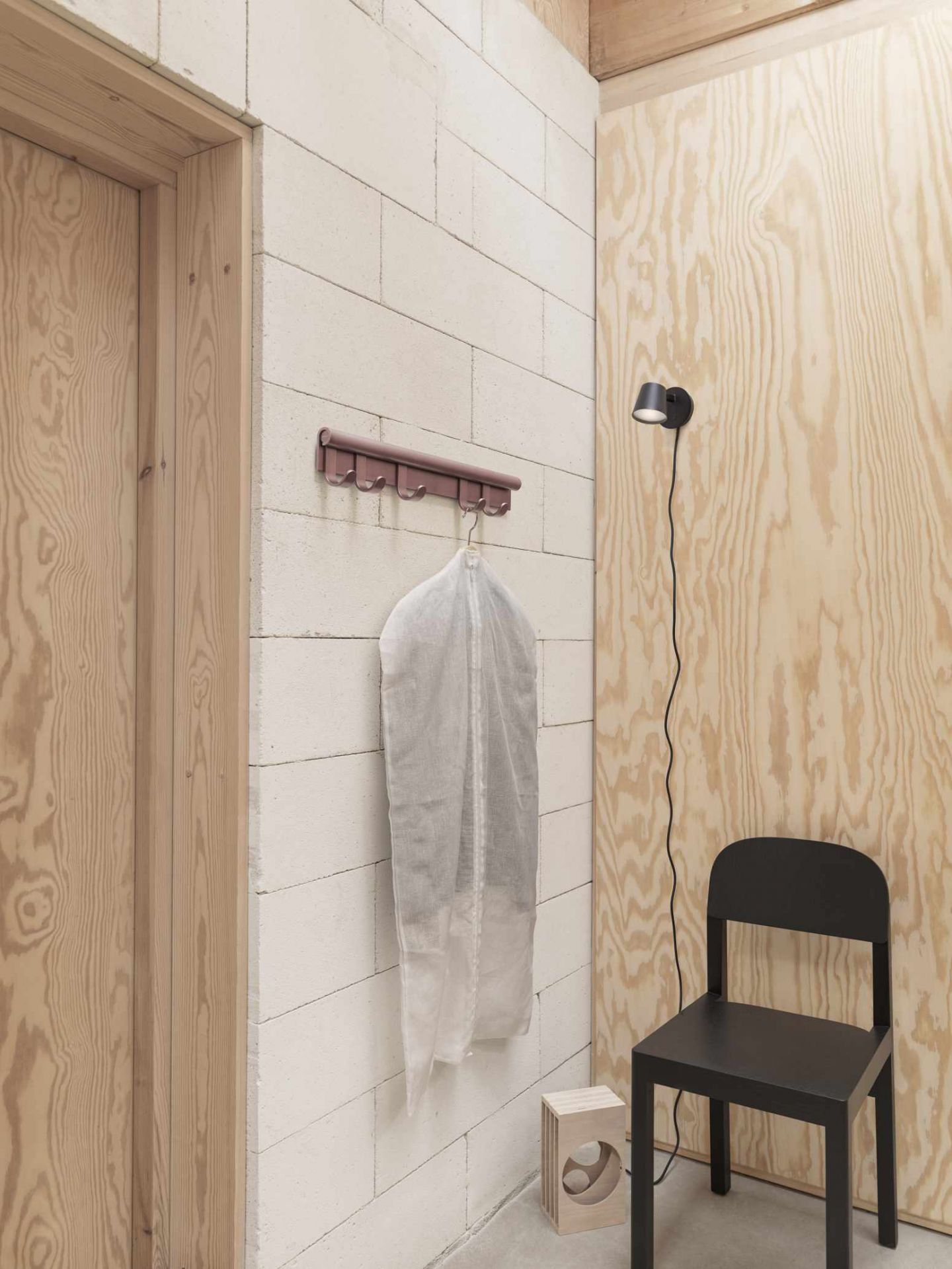 Muuto Coil Coat Rack Wandgarderobe in einem minimalistischen Raum mit Kleidersack, Lampe und Stuhl.