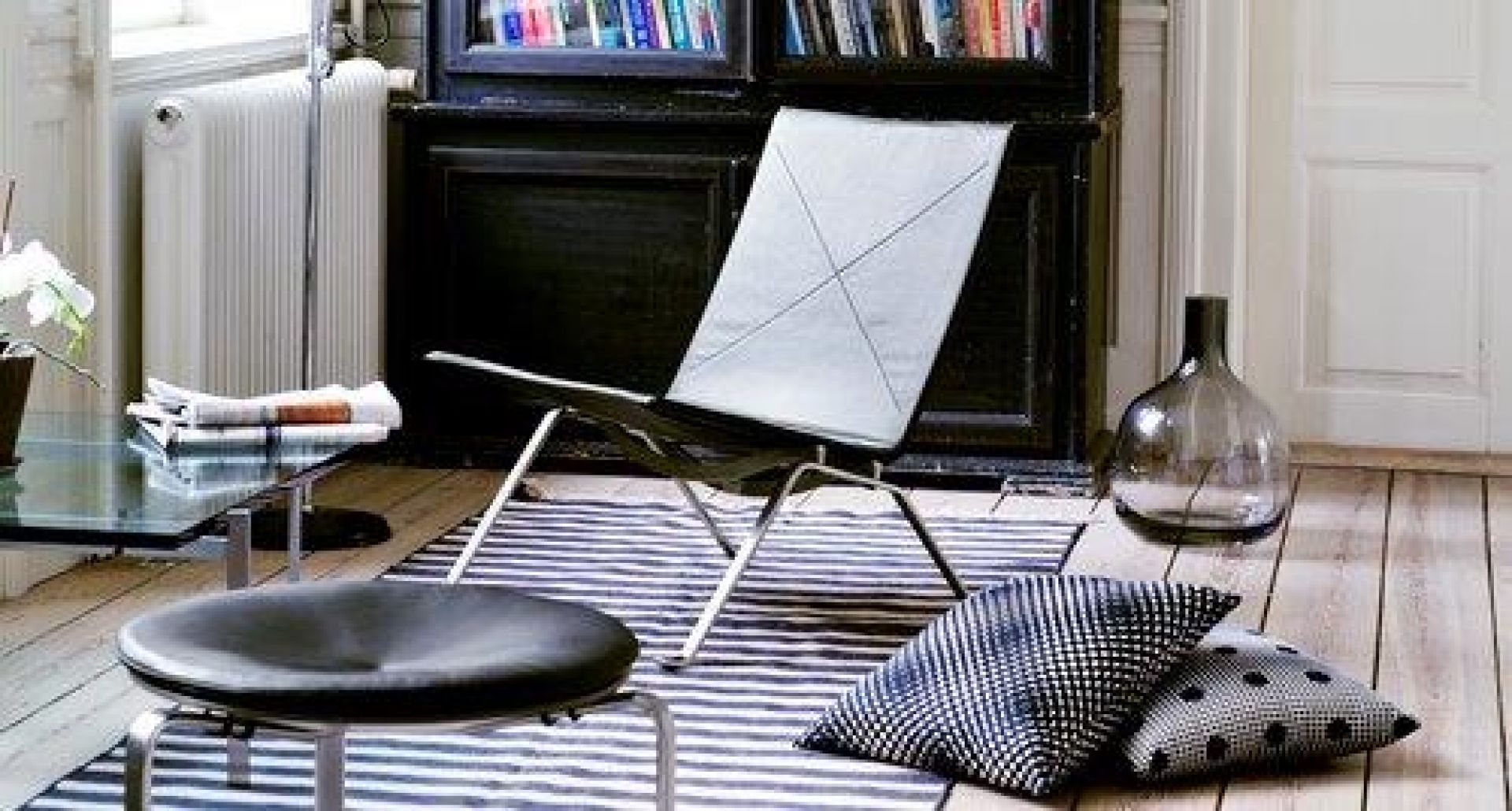 Heller Raum mit Fritz Hansen PK22 Sessel, Hocker, Glasvase und Kissen auf einem gestreiften Teppich.