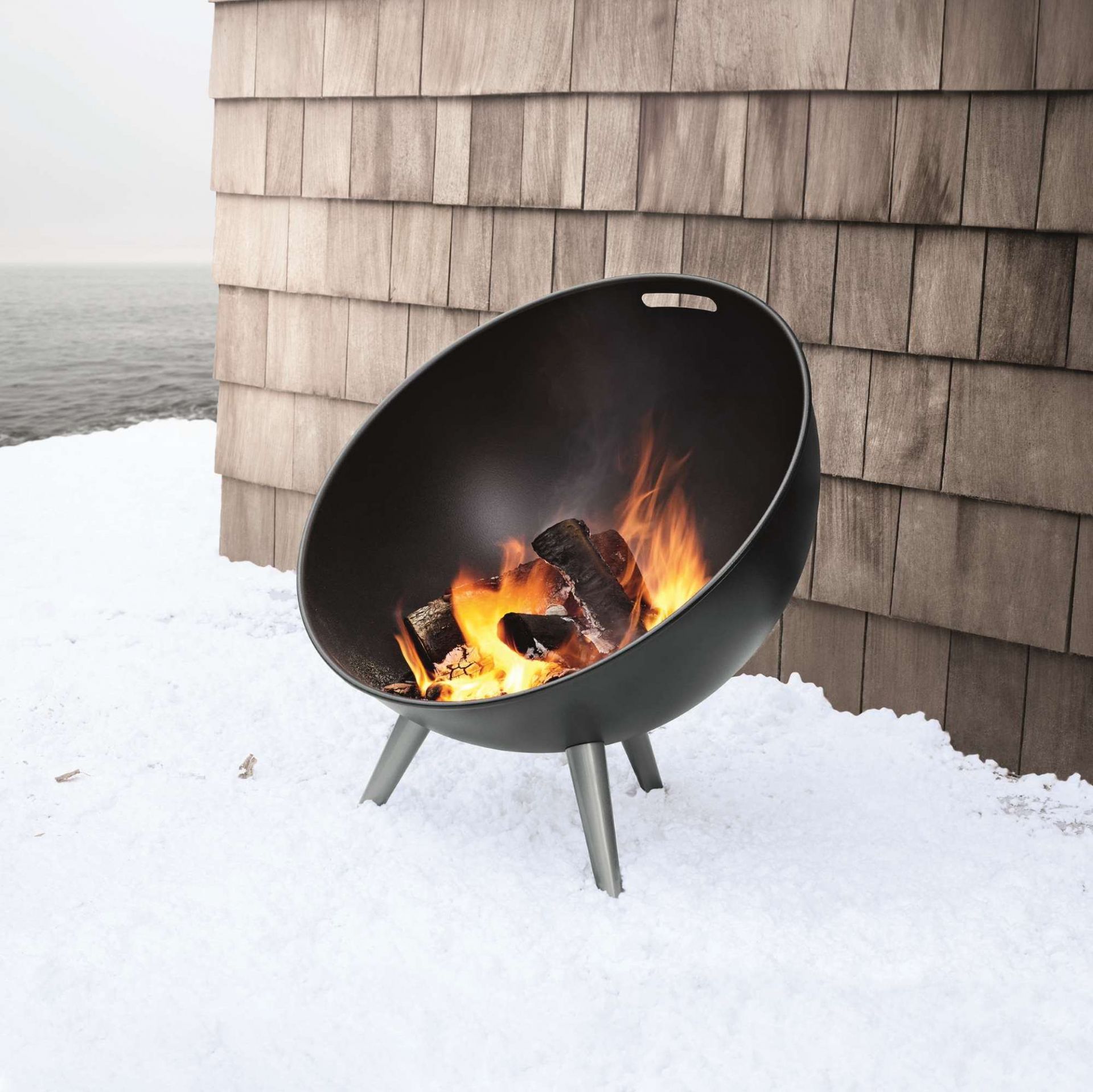 Eva Solo Fire Globe Feuerschale mit brennendem Holz auf Schnee vor einer Holzwand.