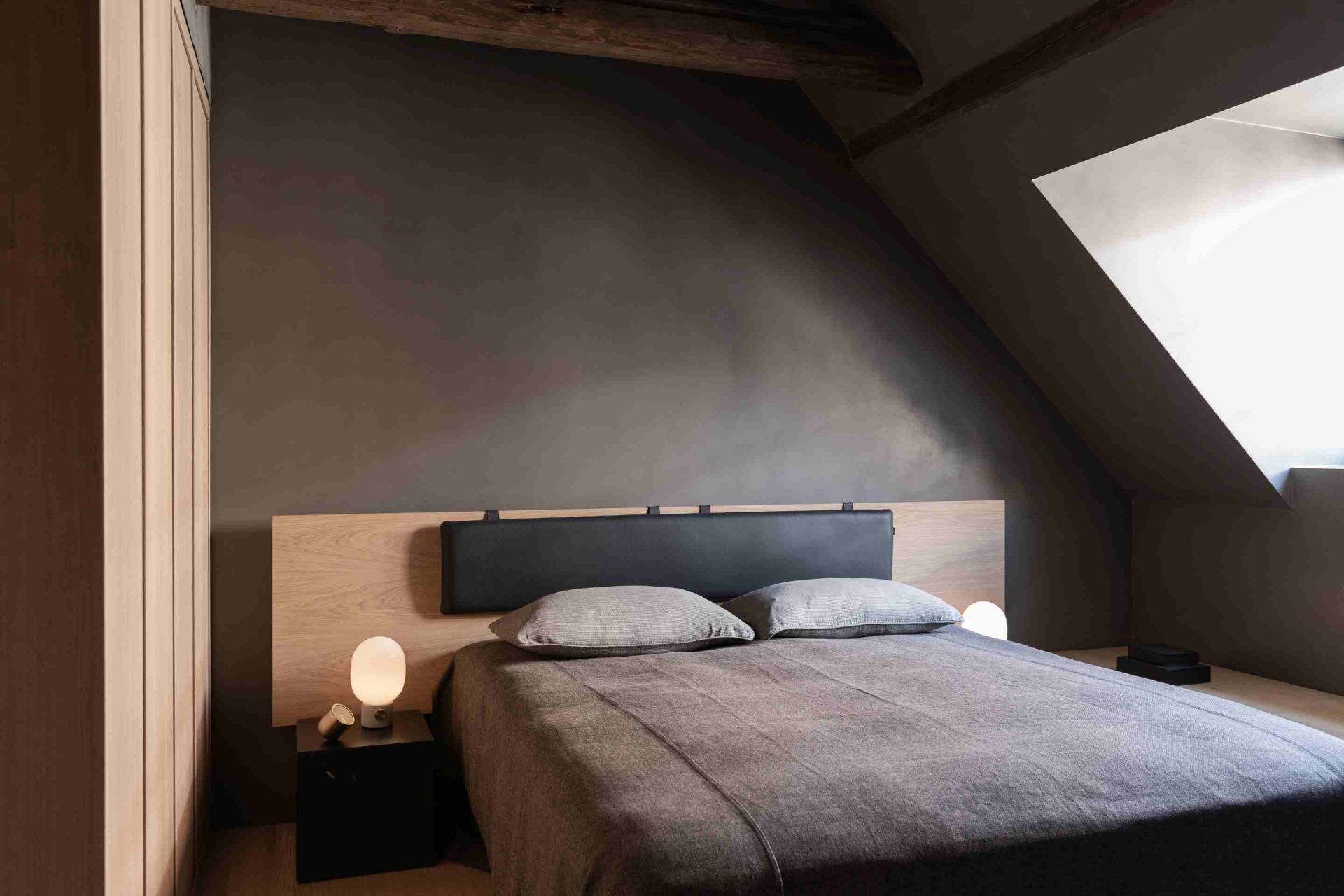 Schlafzimmer mit Audo Plinth Cubic Beistelltisch in Braun und Grau, Nachttischlampe und Bett.