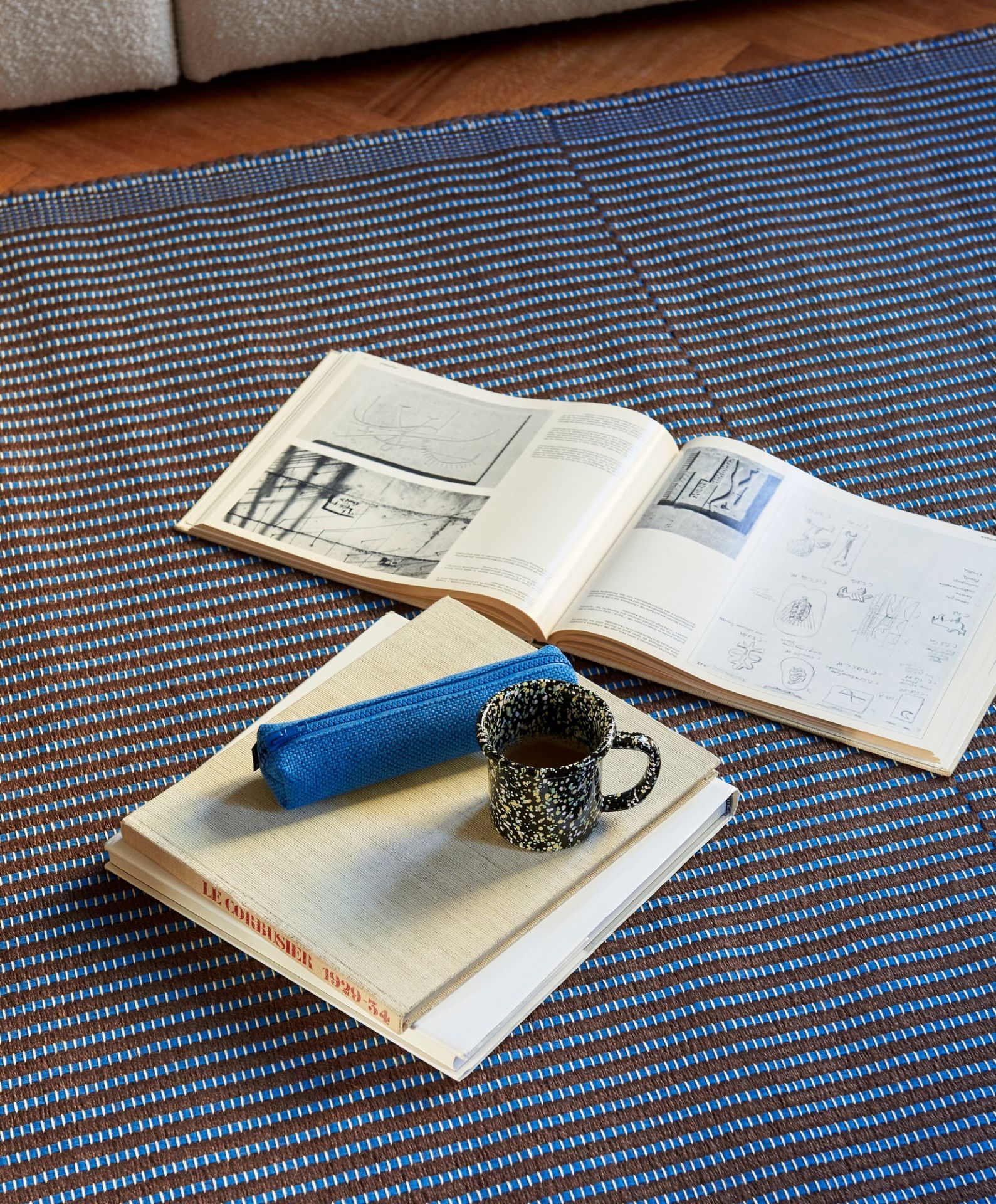 Detailaufnahme des Hay Tapis Teppichs mit blau-braunem Streifenmuster, Büchern, Stifteetui und Kaffeetasse.