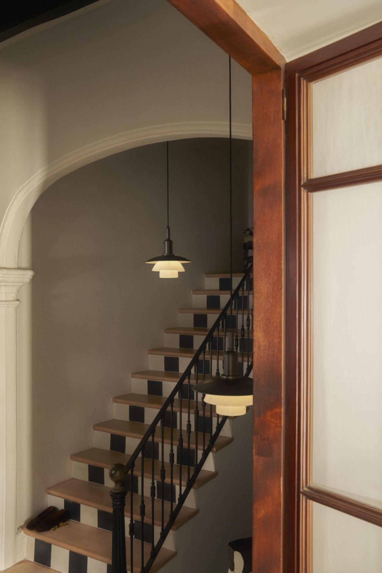 PH 3/3 Pendelleuchte Aged Brass von Louis Poulsen über Treppe mit Schwarz-Weiß-Akzenten, gesehen durch Holztürrahmen.