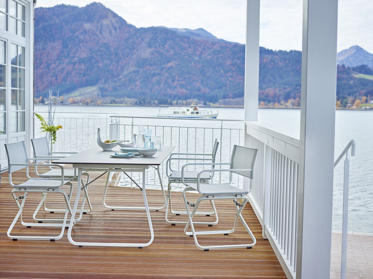 Weishäupl Ahoi Tisch Outdoor mit Stühlen auf einer Terrasse mit Seeblick und Bergen im Hintergrund.